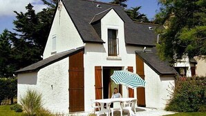 A small, single-story house with a steeply pitched roof, white exterior walls, and wooden shutters. There is a small porch with a table and chairs, and an umbrella.
