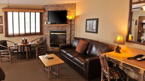 A cozy living room with a fireplace, leather sofa, wooden chairs, a coffee table, and a flat-screen TV mounted on a stone wall. - Boarders Inn & Suites by Cobblestone Hotels – Waukon (Waukon)