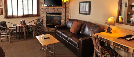 A cozy living room with a fireplace, leather sofa, wooden chairs, a coffee table, and a flat-screen TV mounted on a stone wall.