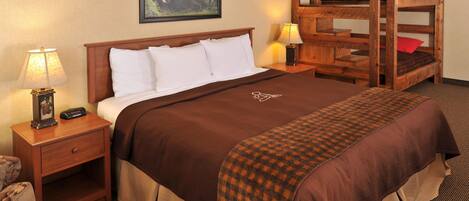A hotel room with a wooden bunk bed, a single bed with a brown bedspread, a nightstand with a lamp, and a framed picture on the wall.