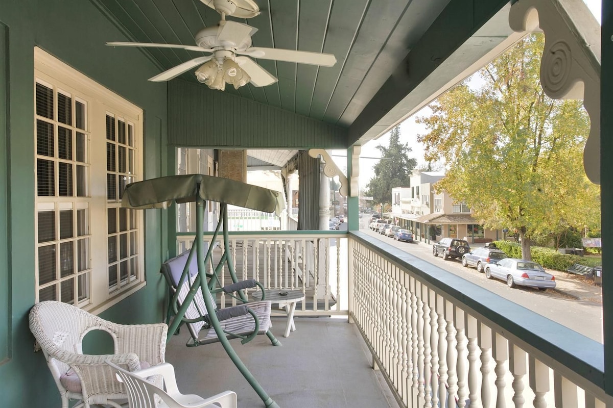 A porch with a ceiling fan, wicker chairs, a swing, and a view of a street with parked cars.