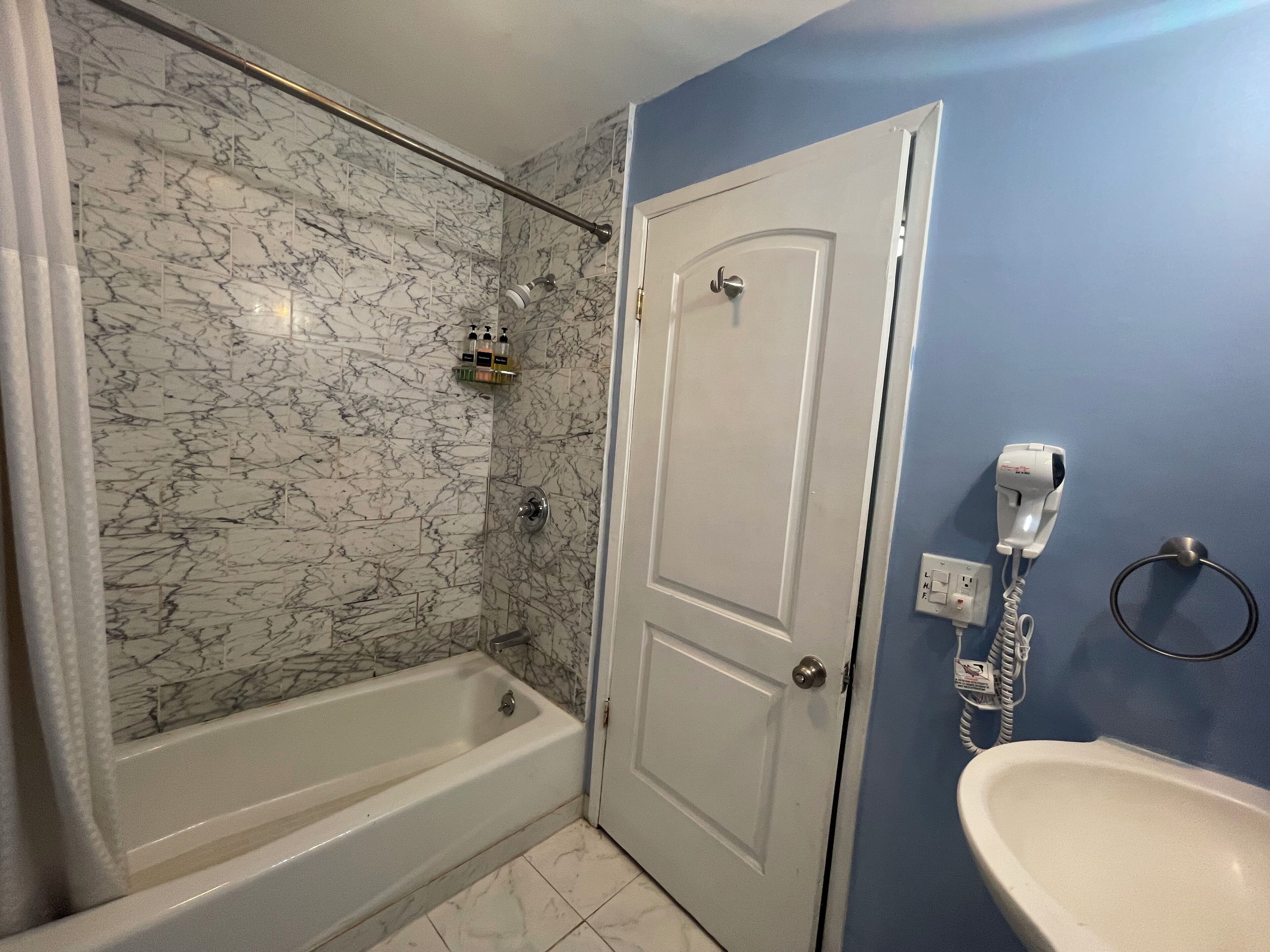 Une salle de bain avec un mur de douche en marbre, une baignoire blanche, une porte, un sèche-cheveux et un lavabo.