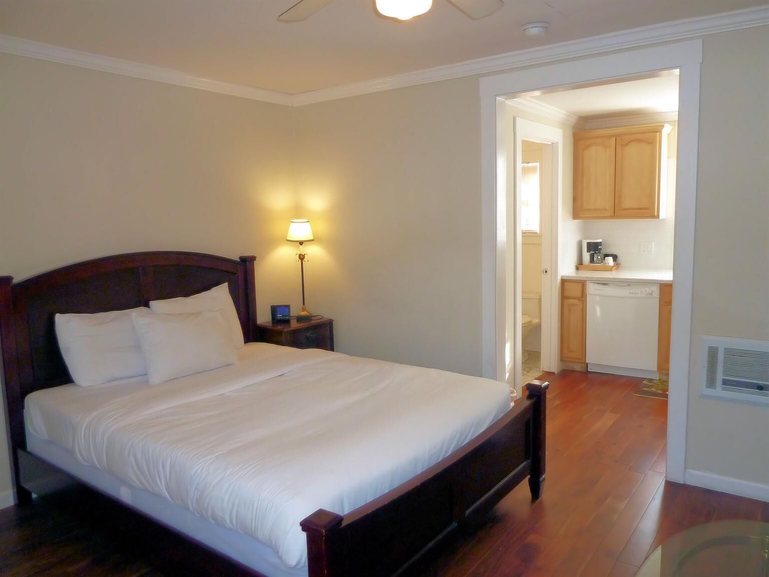 A bedroom with a wooden bed, bedside lamp, and a view into a kitchen area.