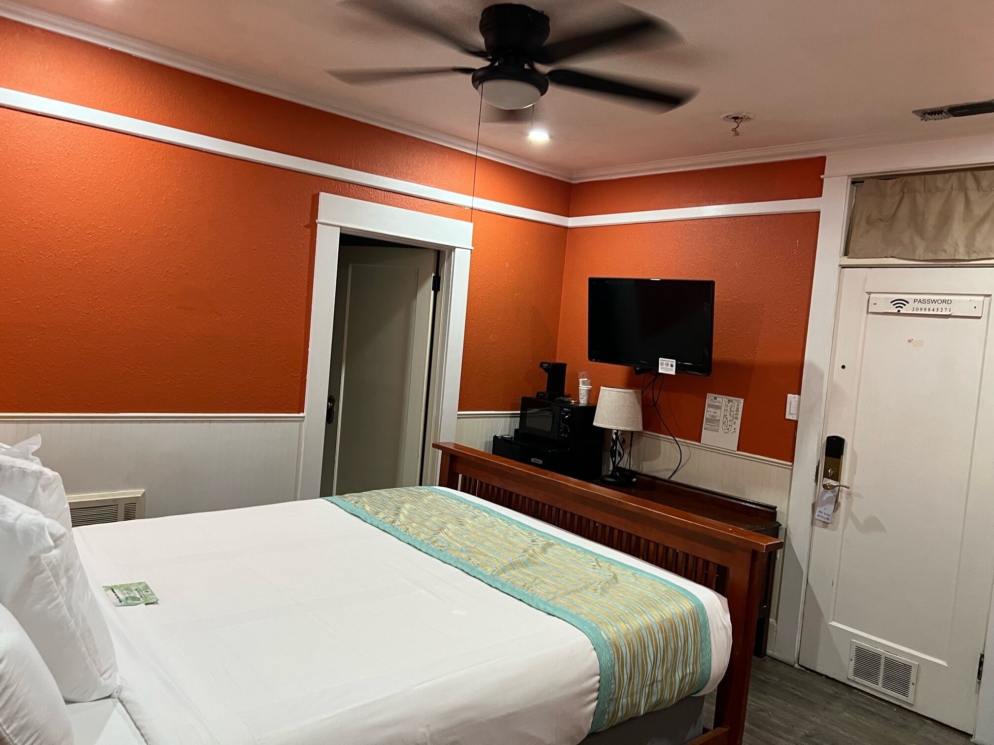 A hotel room with a bed, a ceiling fan, a television, a desk with a lamp, and a door with a sign.