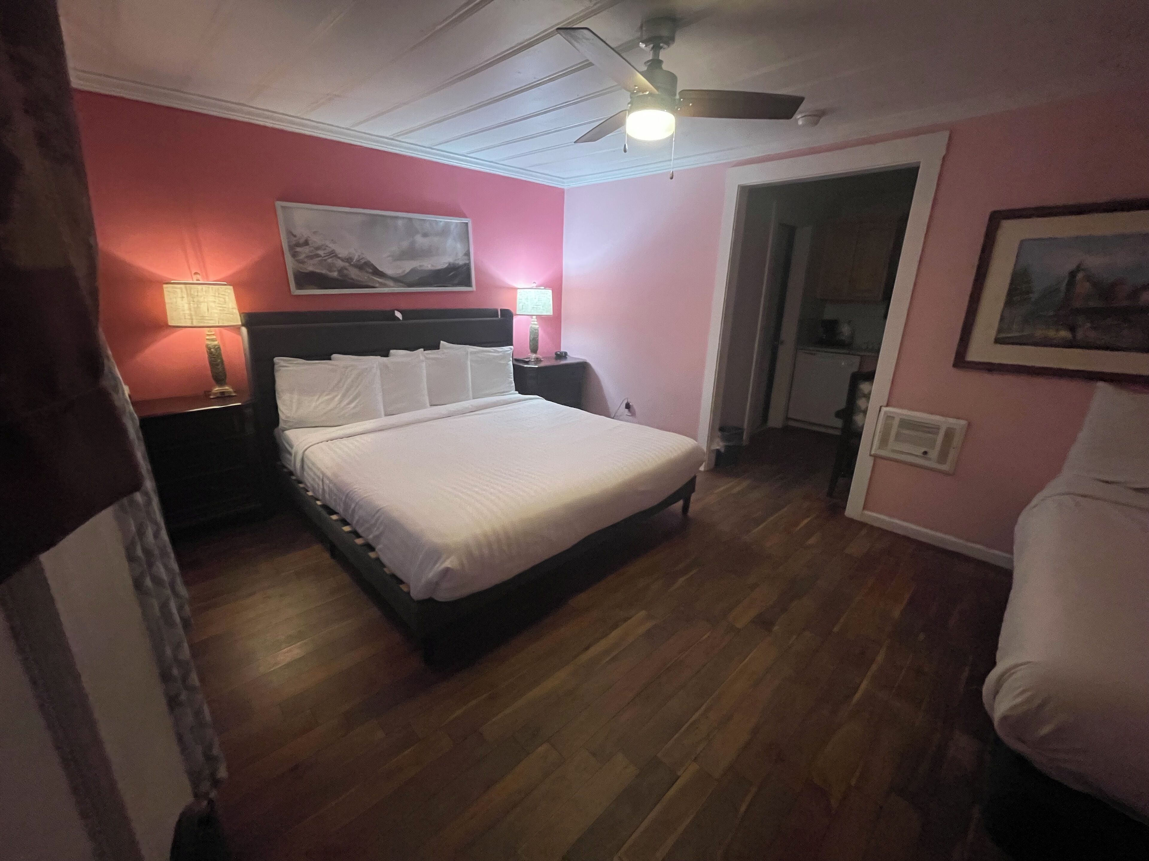 Un dormitorio con cama, mesitas de noche, ventilador de techo y puerta que da al baño.