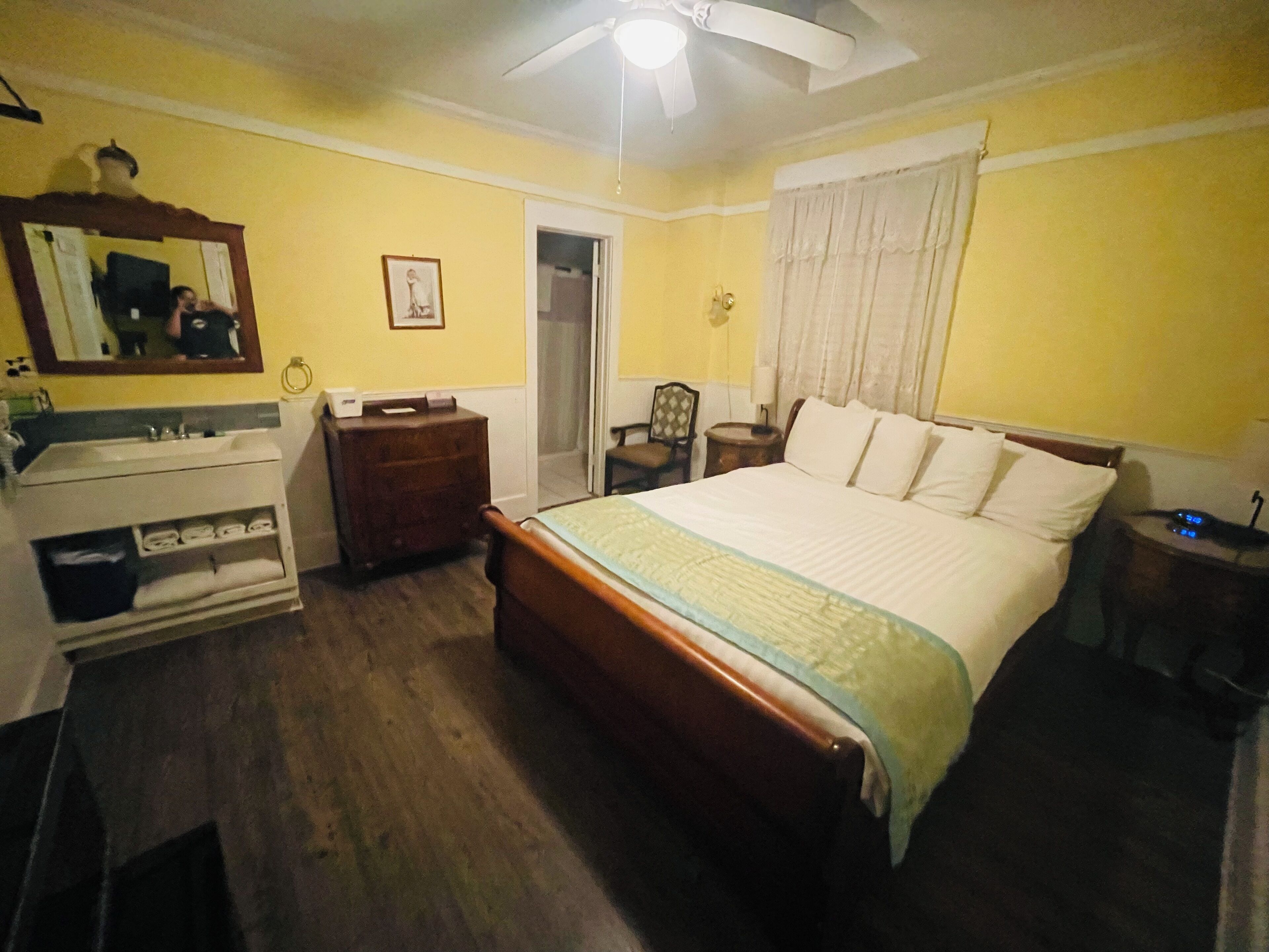 Un dormitorio con cama, cómoda, tocador, espejo, ventilador de techo y ventana con cortinas.