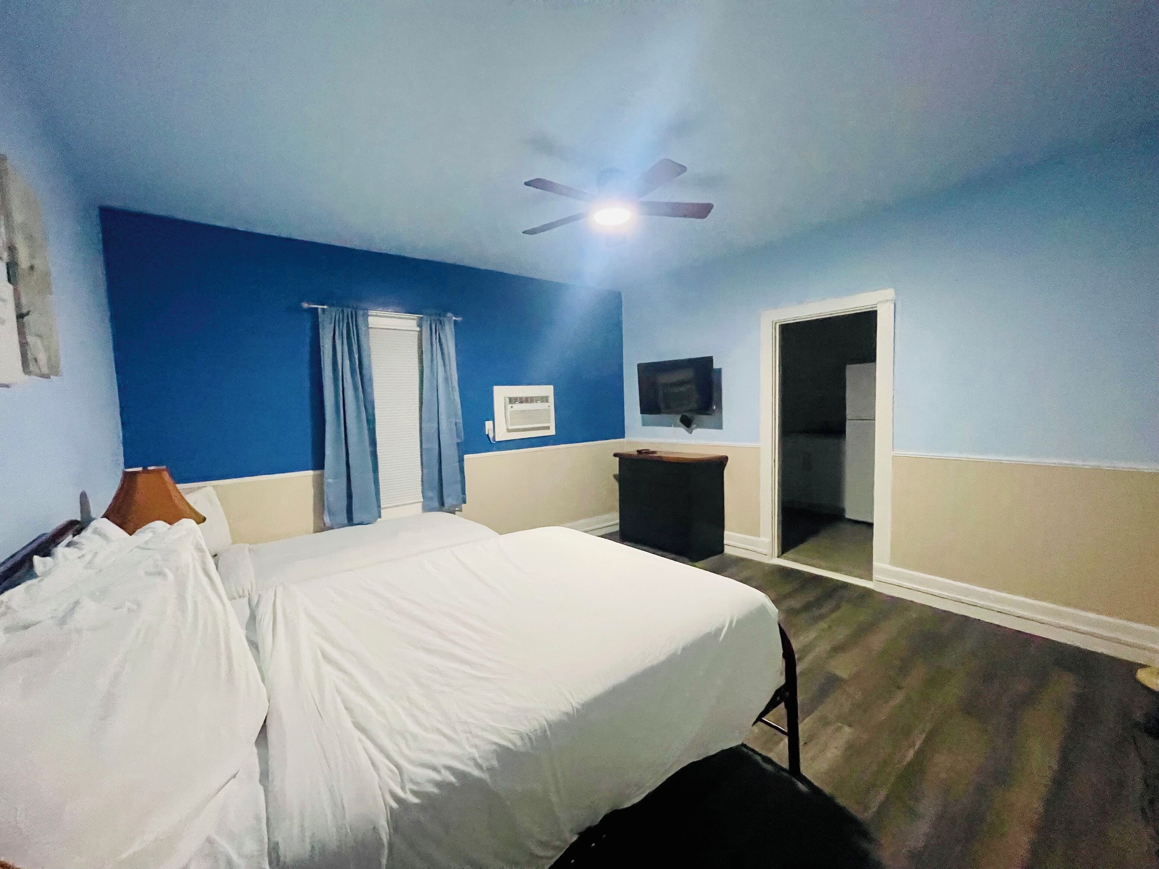 A hotel room with a bed, a desk, a chair, a ceiling fan, a window with curtains, and a television on the wall.