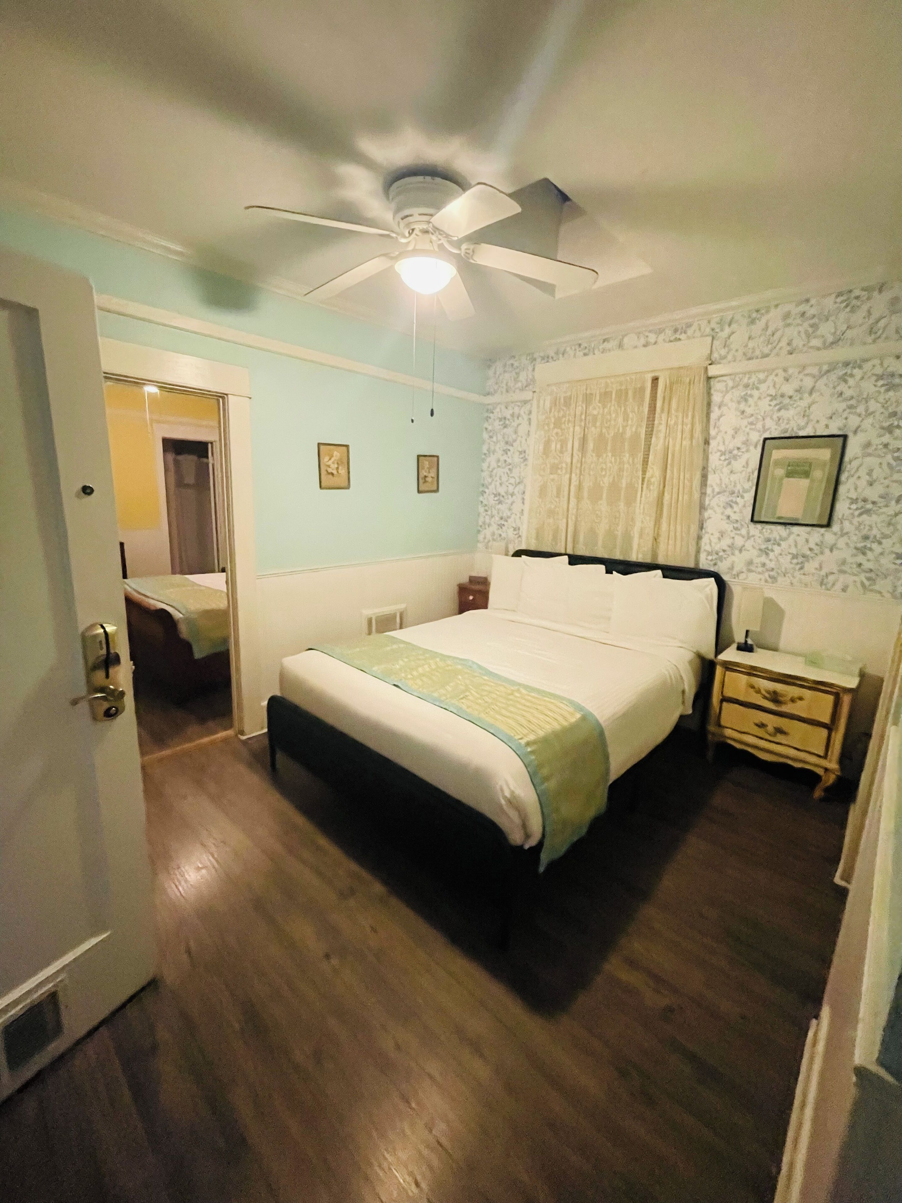 Ein Schlafzimmer mit Bett, Nachttisch, Deckenventilator und einer Tür zu einem anderen Raum.