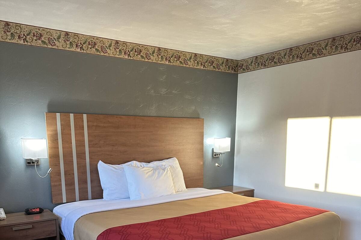 A hotel room with a large bed, a wooden headboard, a nightstand with a lamp, and a window with blinds.