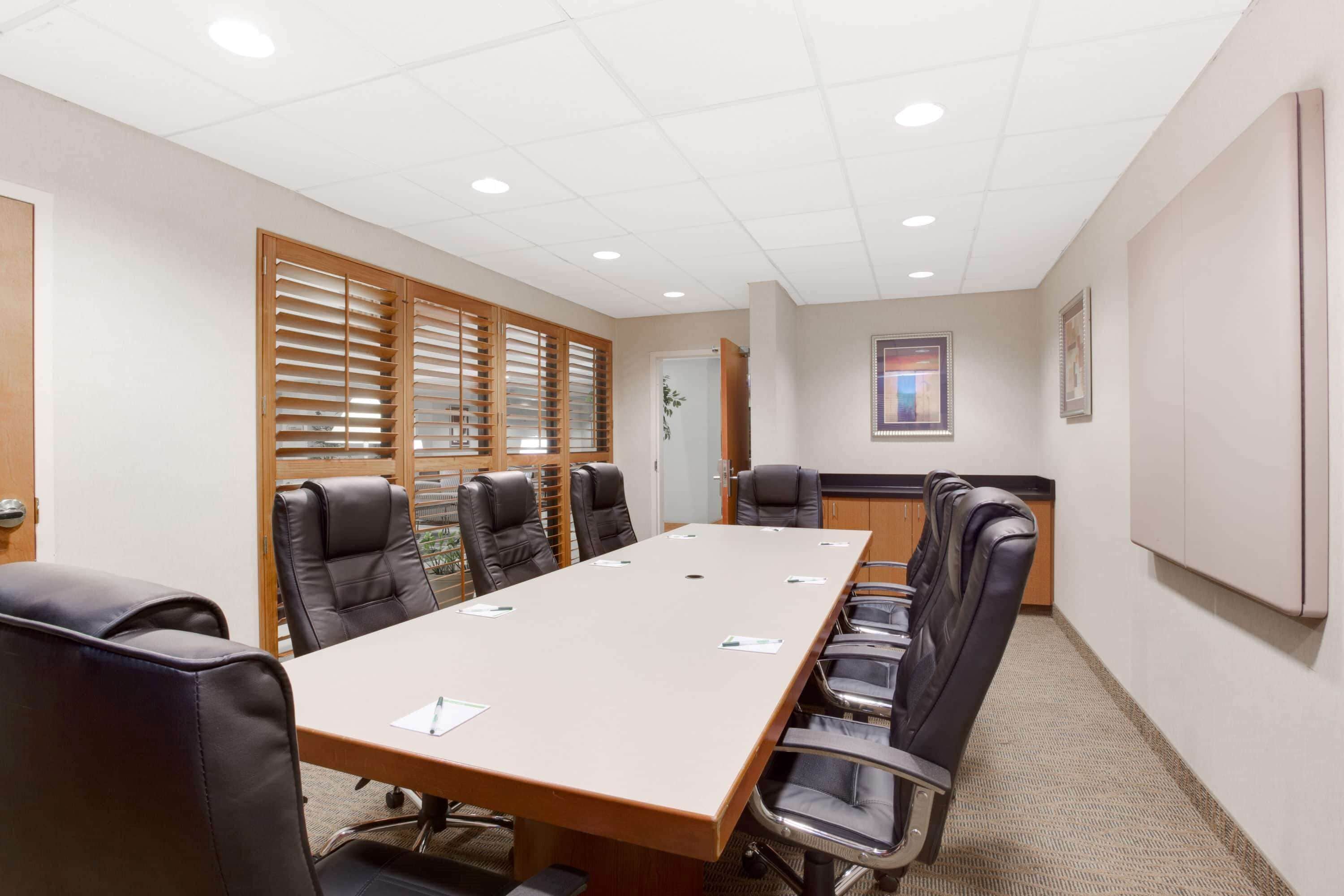 A conference room with a long table, black leather chairs, wooden blinds, and a whiteboard on the wall.