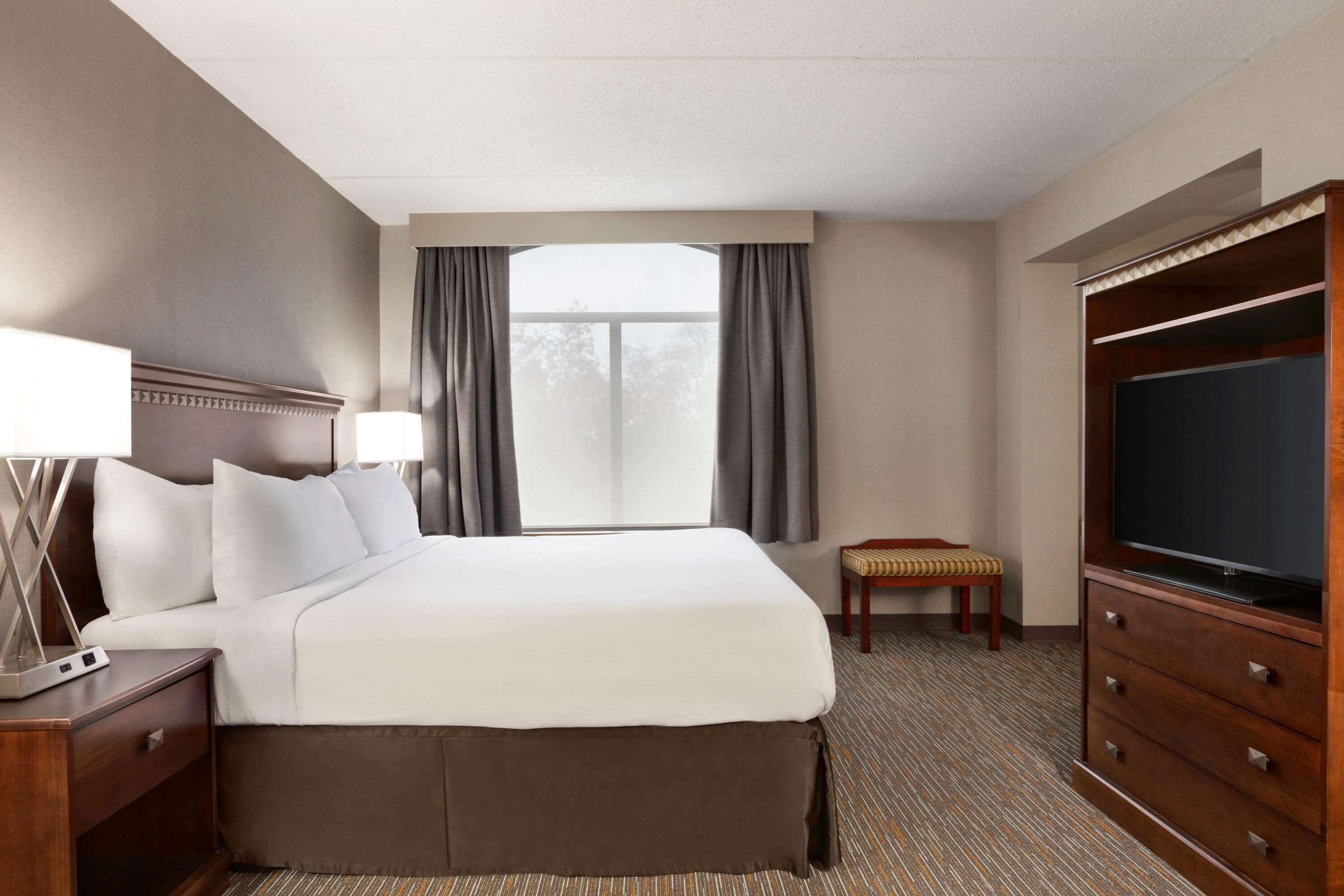 A hotel room with a large bed, a TV, a desk with a lamp, and a window with curtains.
