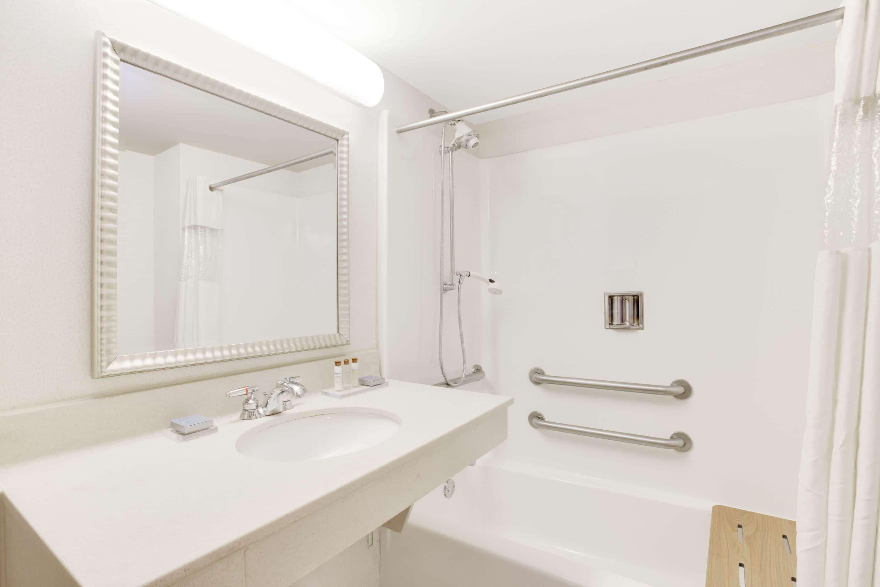 A bathroom with a white sink, mirror, and bathtub. There are handrails on the walls and a showerhead above the bathtub.