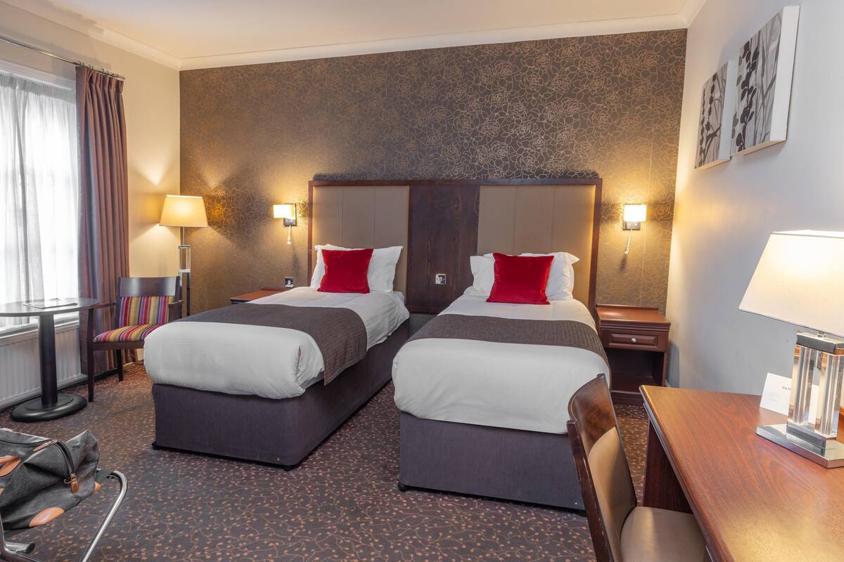 A hotel room with two beds, a desk, a chair, a lamp, and a view of the city.