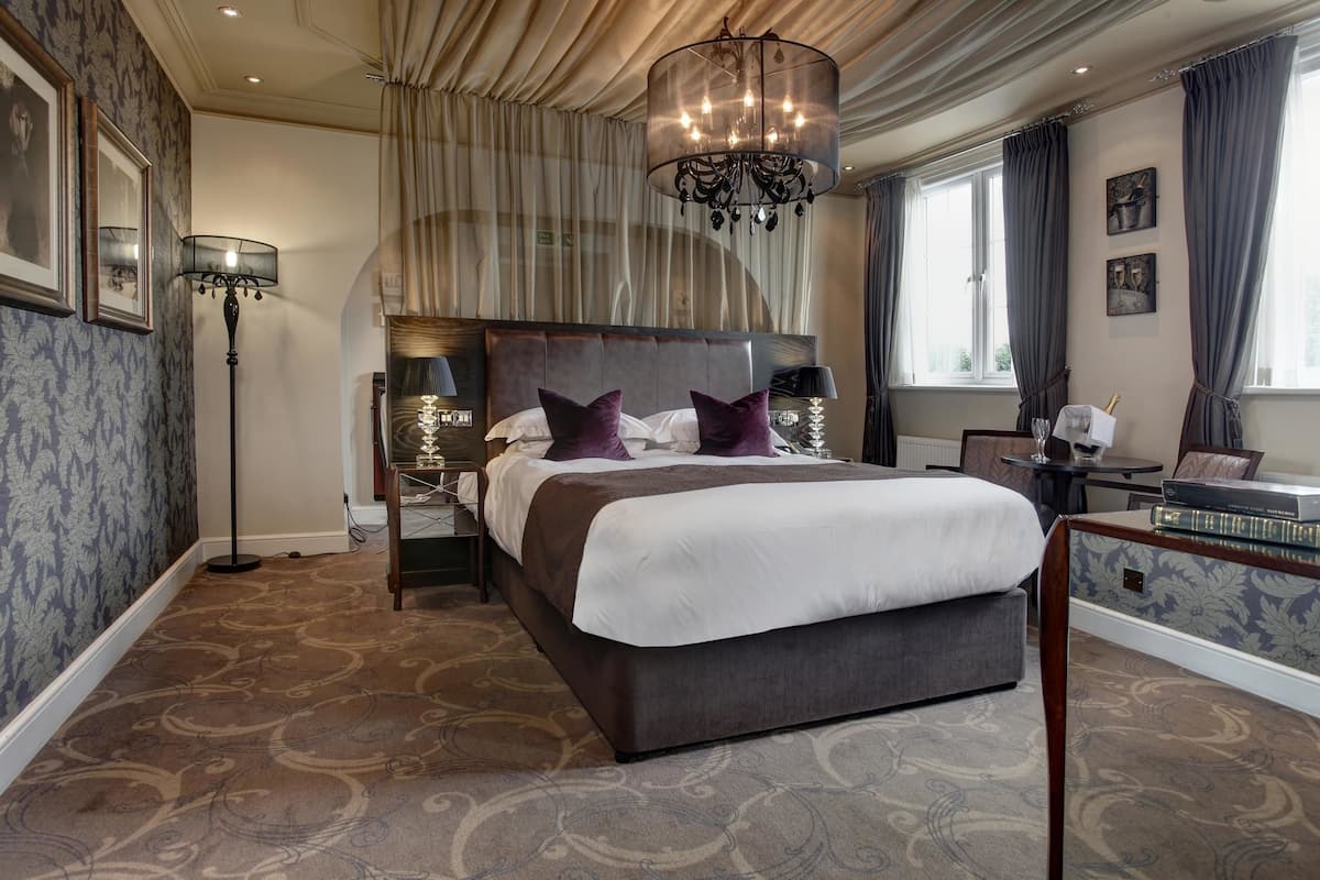 A large bed with a headboard, a floor lamp, and a table with books and a glass of wine.