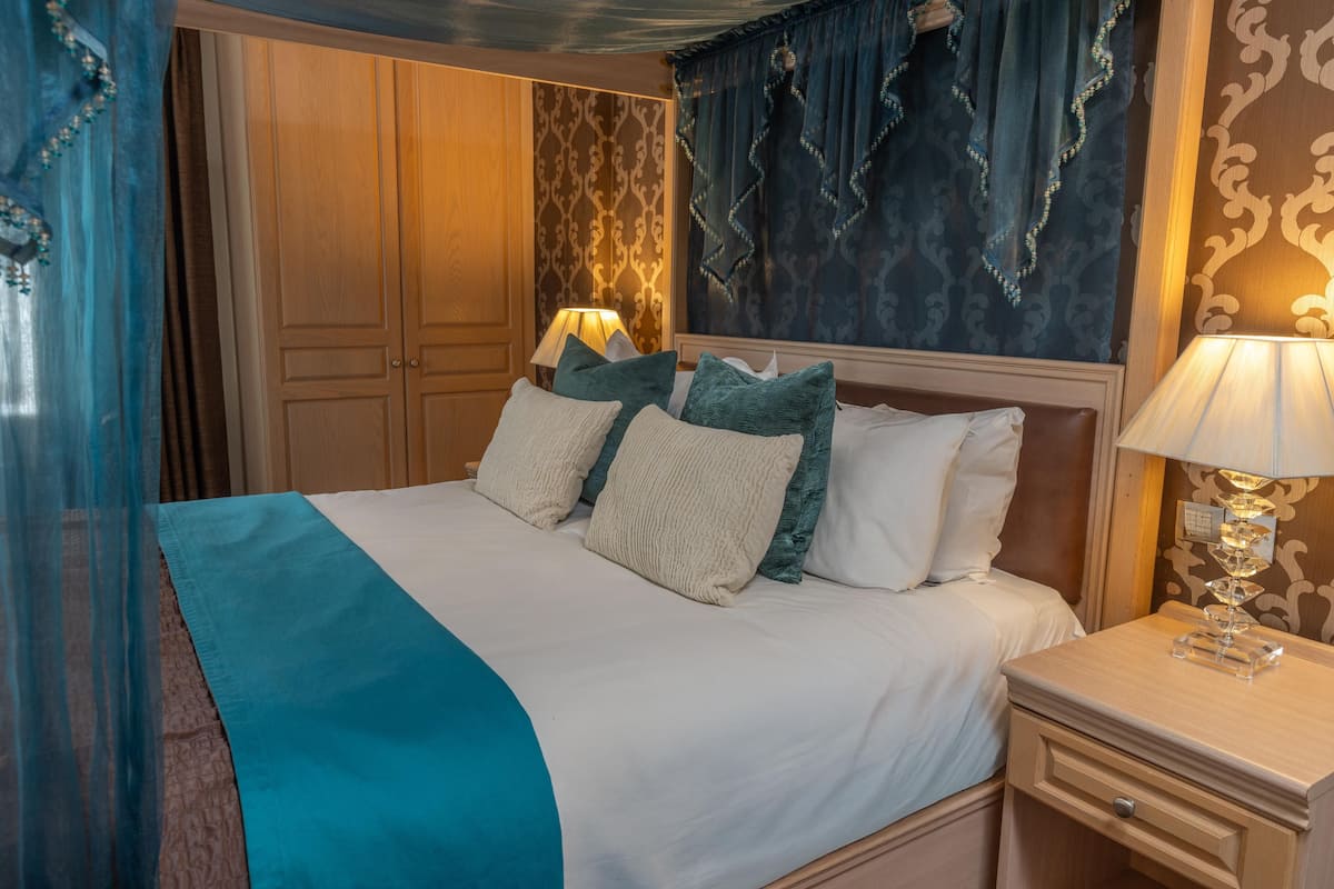 A neatly made bed with white and teal bedding, a wooden nightstand with a lamp, and a patterned wall behind the bed.
