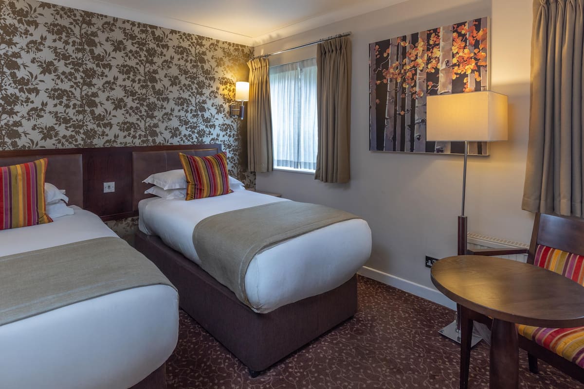 A hotel room with two beds, a small table, a lamp, and a painting on the wall.