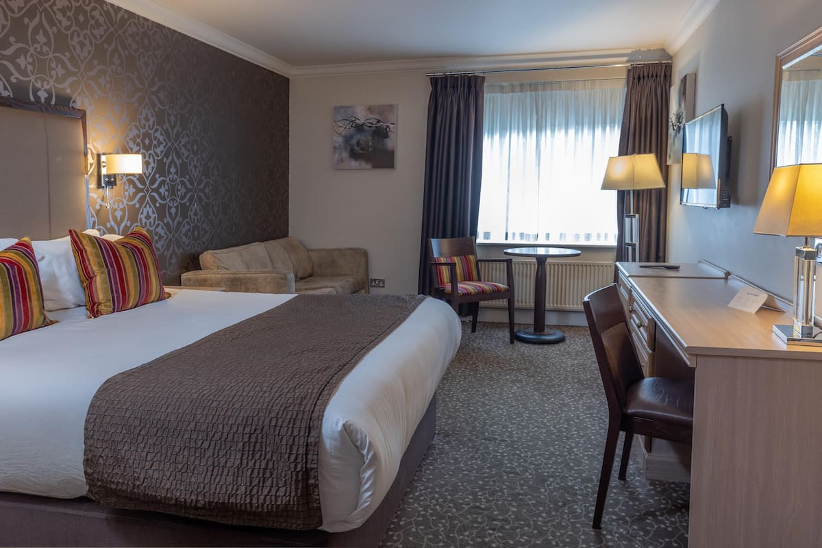 A hotel room with a large bed, a desk with a chair, a small table, and a window with curtains.