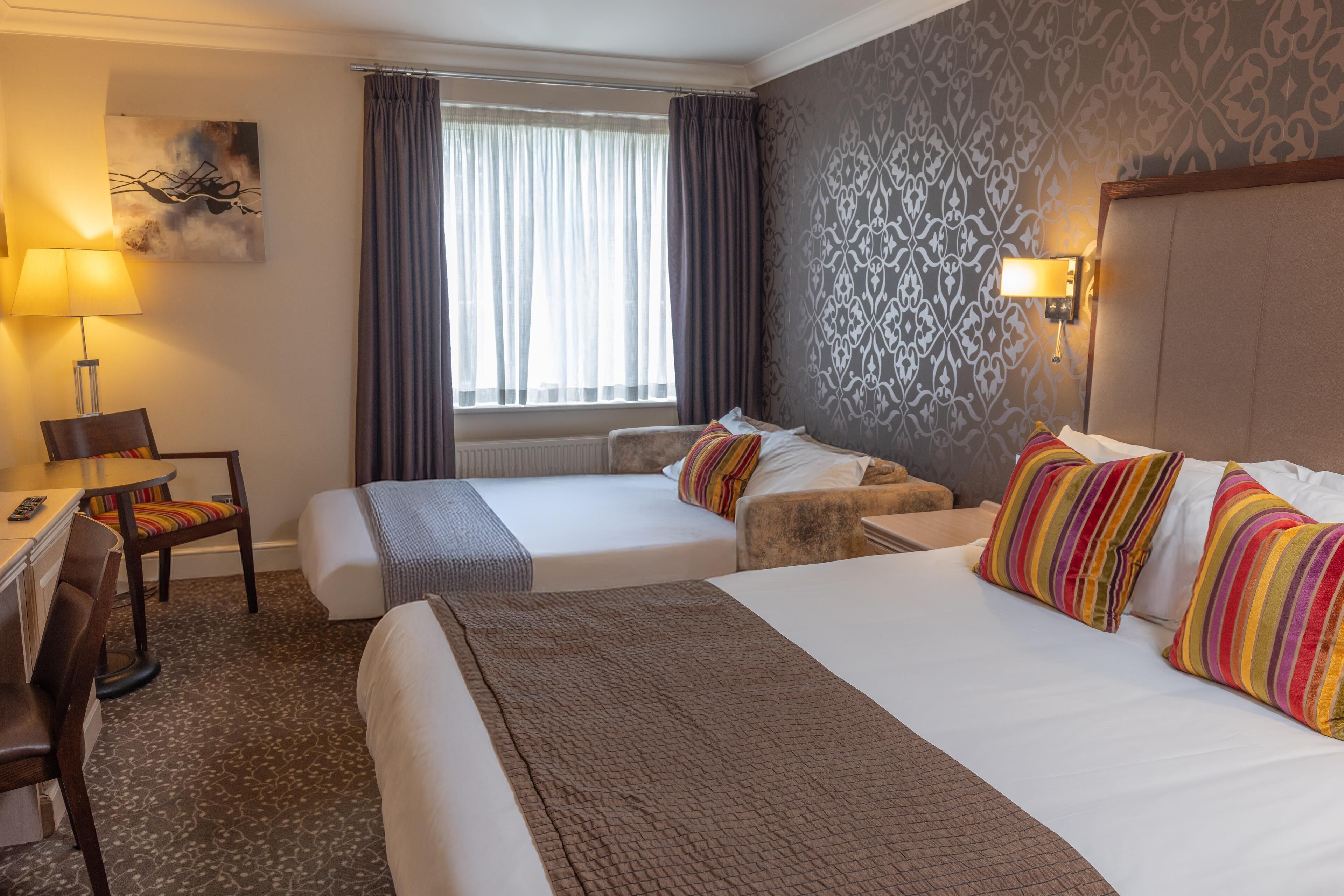 Une chambre d’hôtel avec deux lits, un bureau, une chaise, une lampe et une fenêtre avec des rideaux.