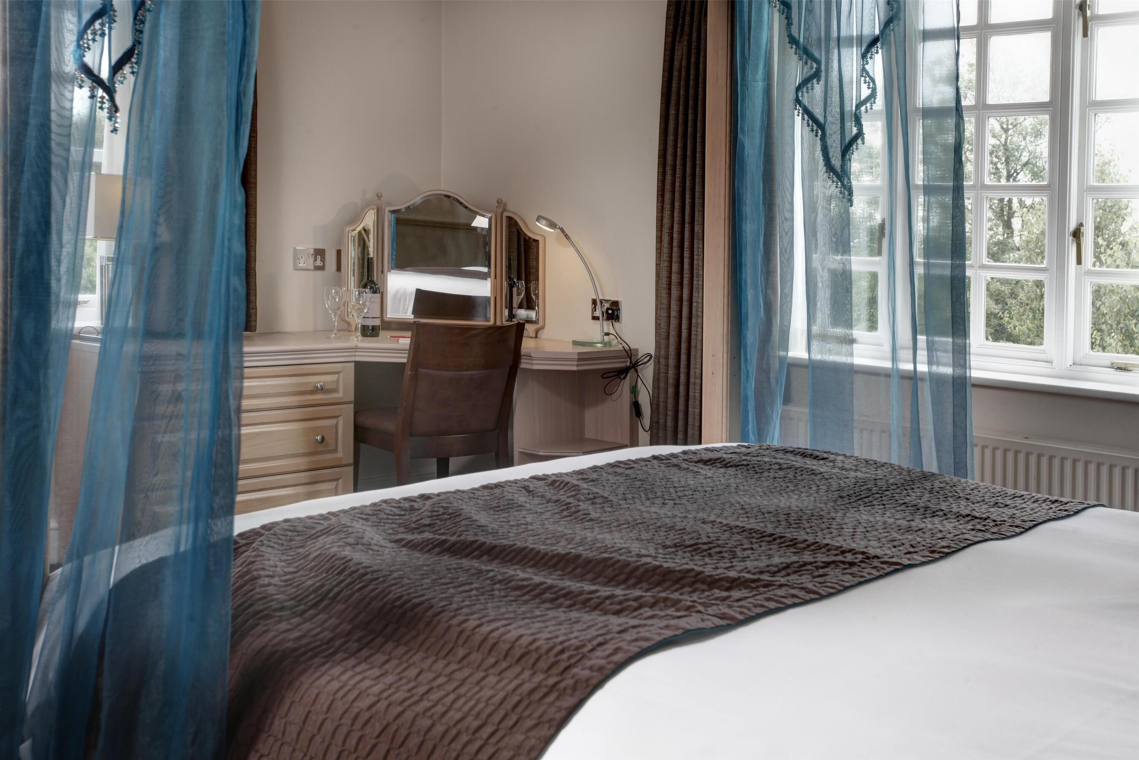 A bedroom with a bed, a desk, a chair, a mirror, and a window with curtains.