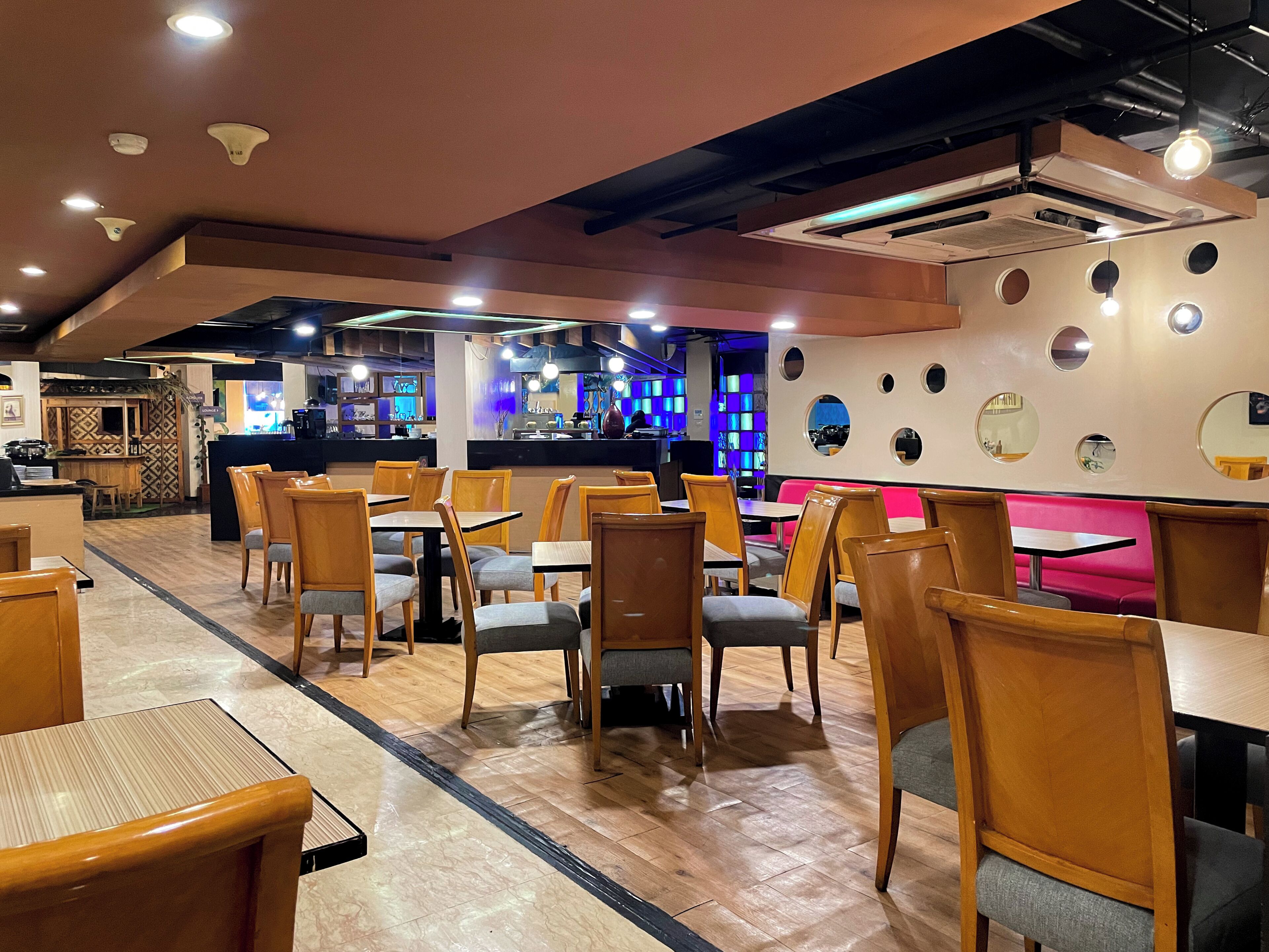 A restaurant interior with wooden tables and chairs, a bar area, and a wall with circular cutouts.