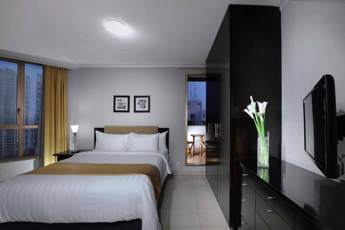 A hotel room with a bed, bedside lamps, a TV, and a view of the cityscape through the window.