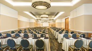 Un salón de conferencias con filas de mesas y sillas, equipado con micrófonos y vasos, bajo una lámpara moderna.
