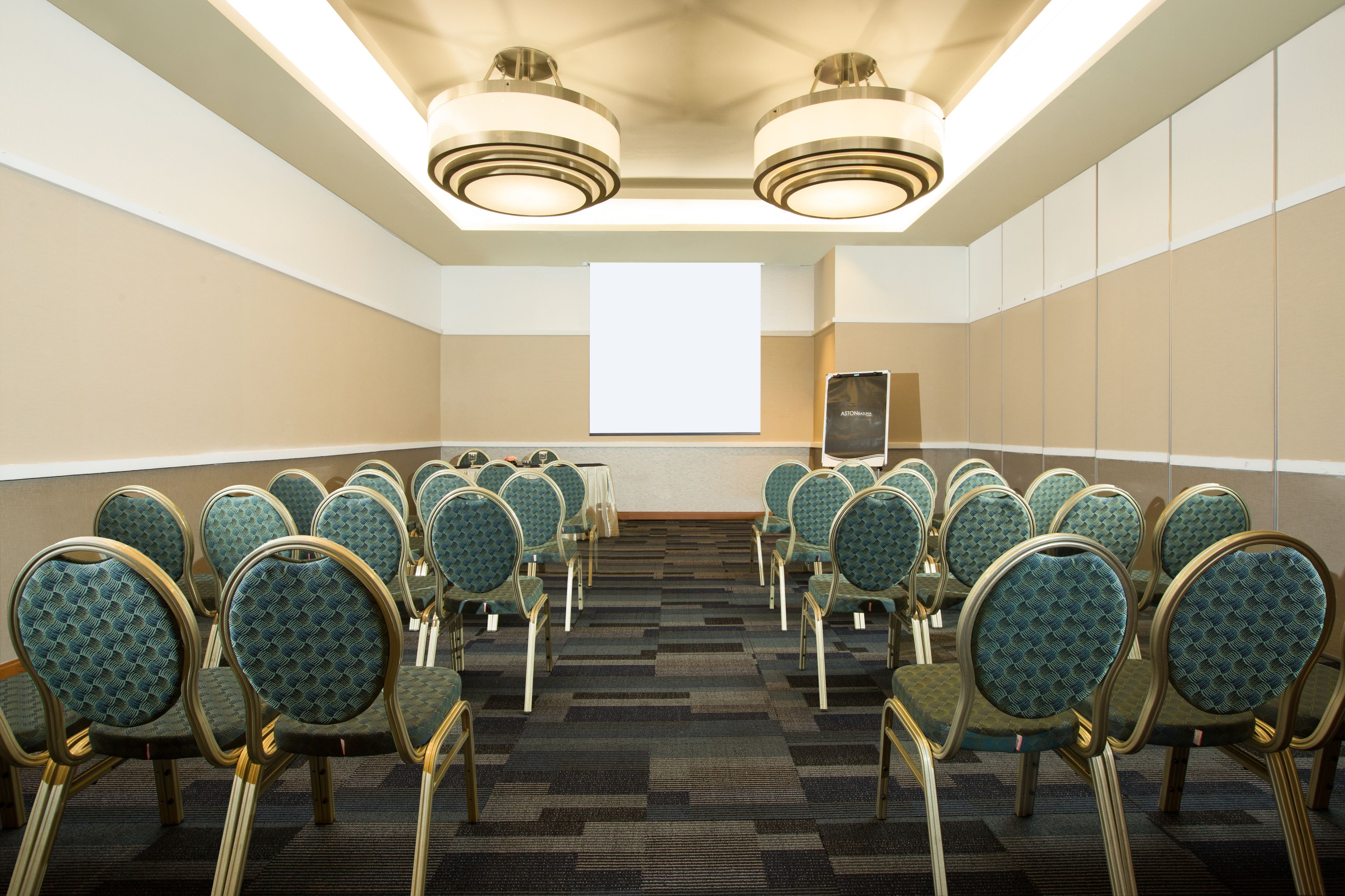 A conference room with rows of green upholstered chairs, a carpeted floor, and a large projection screen at the front.