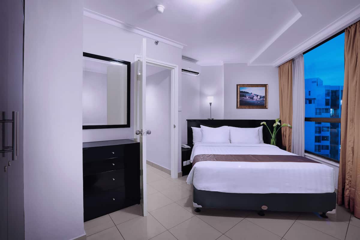A hotel room with a large bed, a dresser, a nightstand, a lamp, a framed picture on the wall, and a view of buildings through a window.