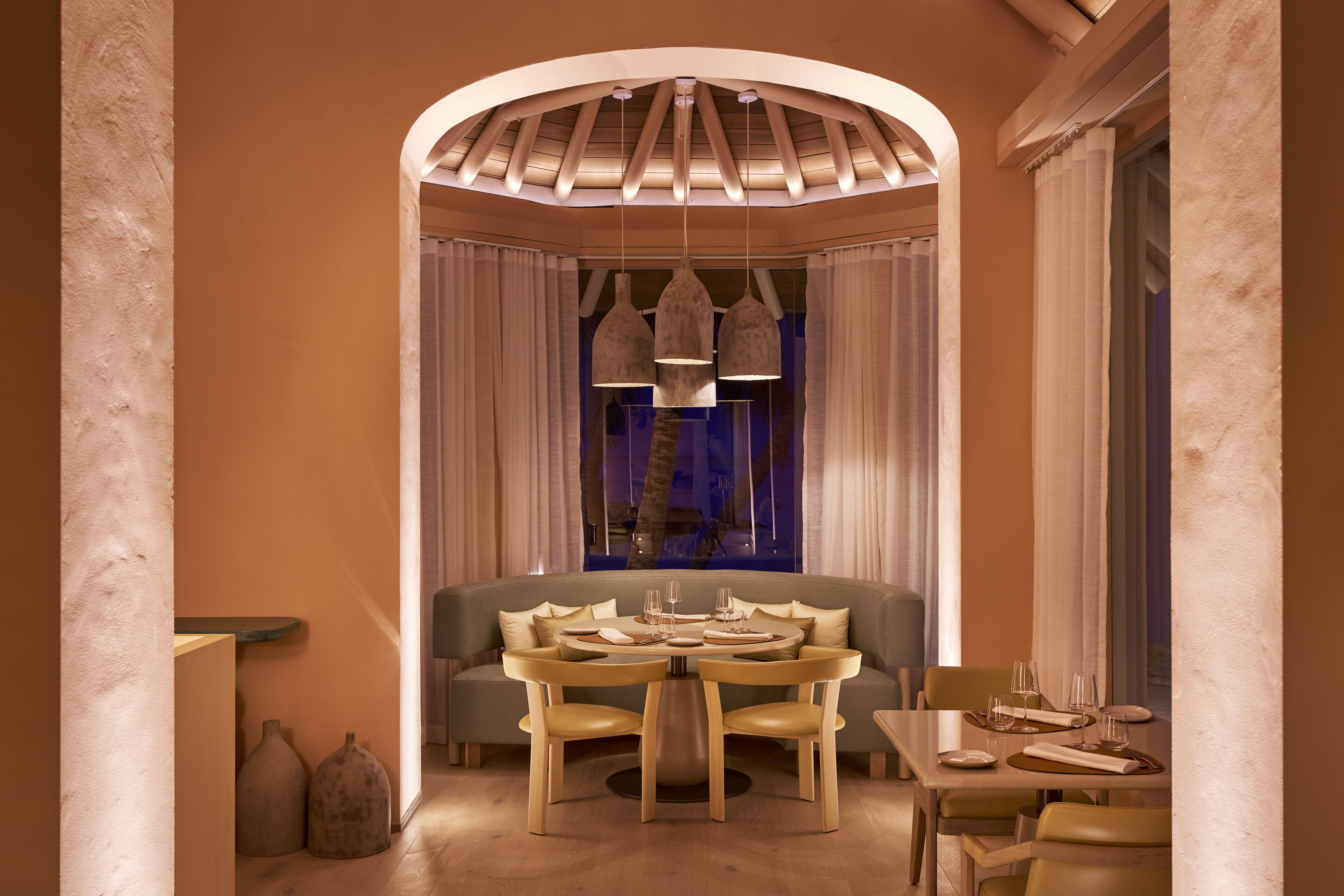 A dining area with a round table, chairs, and a sofa. There is a chandelier above the table and a window with curtains behind the sofa.