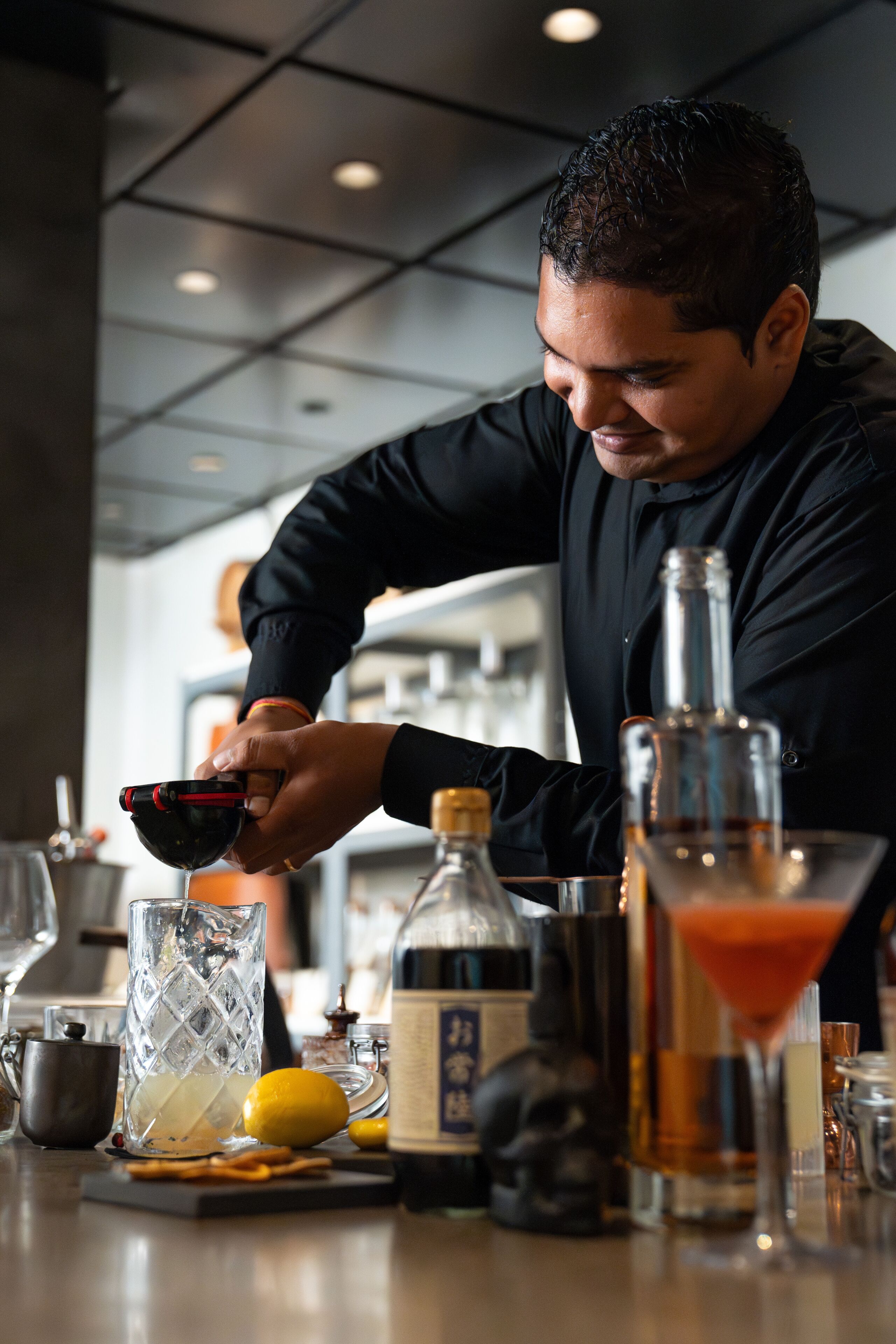 Un bartender preparando una bebida con un agitador, una copa de martini y una botella de licor sobre una barra.
