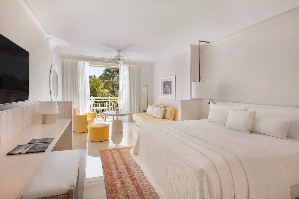 A modern hotel room with a large bed, a desk, and a balcony with a view of palm trees.