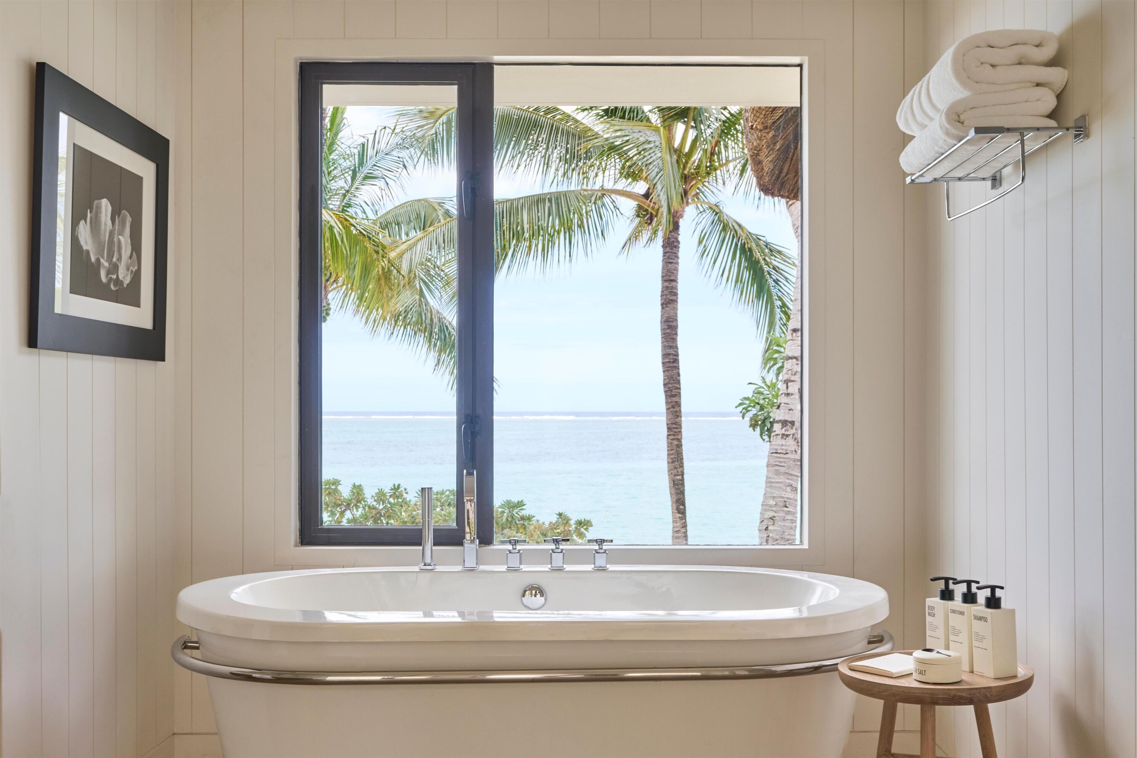 Een badkamer met een bad, een raam met uitzicht op palmbomen en de zee, een handdoekrek en een klein houten krukje met toiletartikelen.