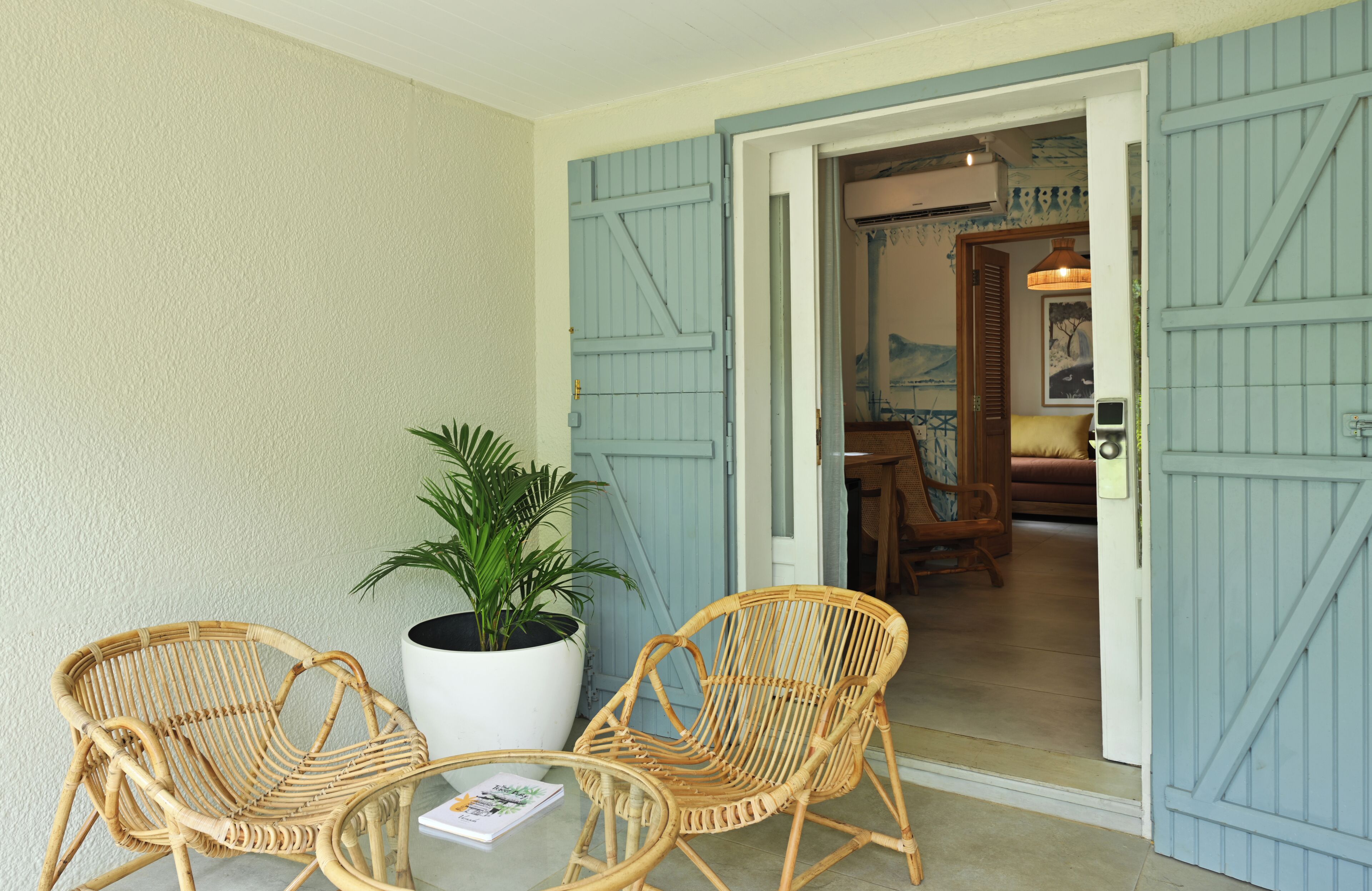 A room with two wicker chairs and a table, a potted plant, and a door leading to another room with a bed and a painting.
