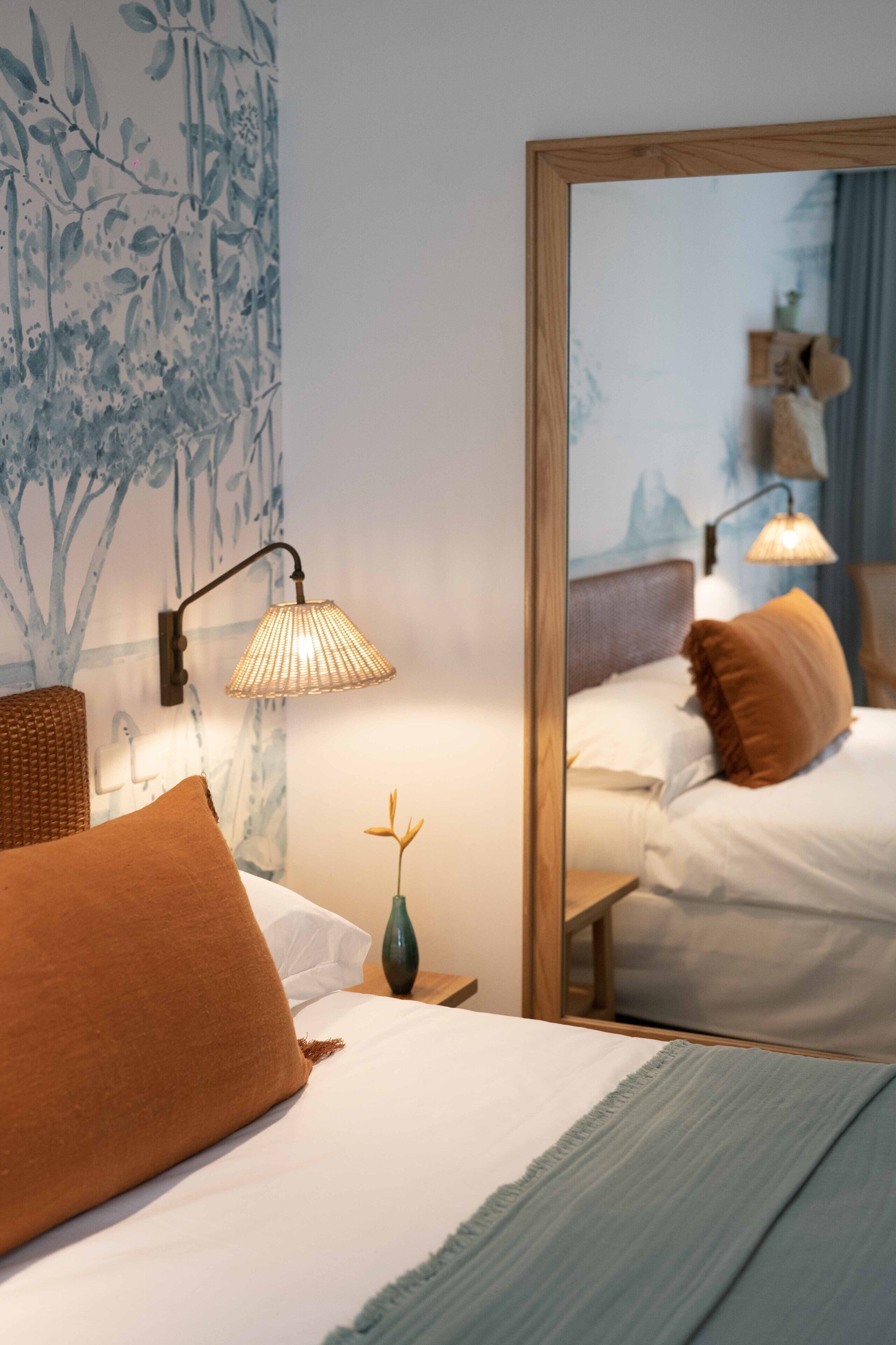 A neatly made bed with a brown pillow, a bedside lamp, and a wall-mounted mirror reflecting the room.