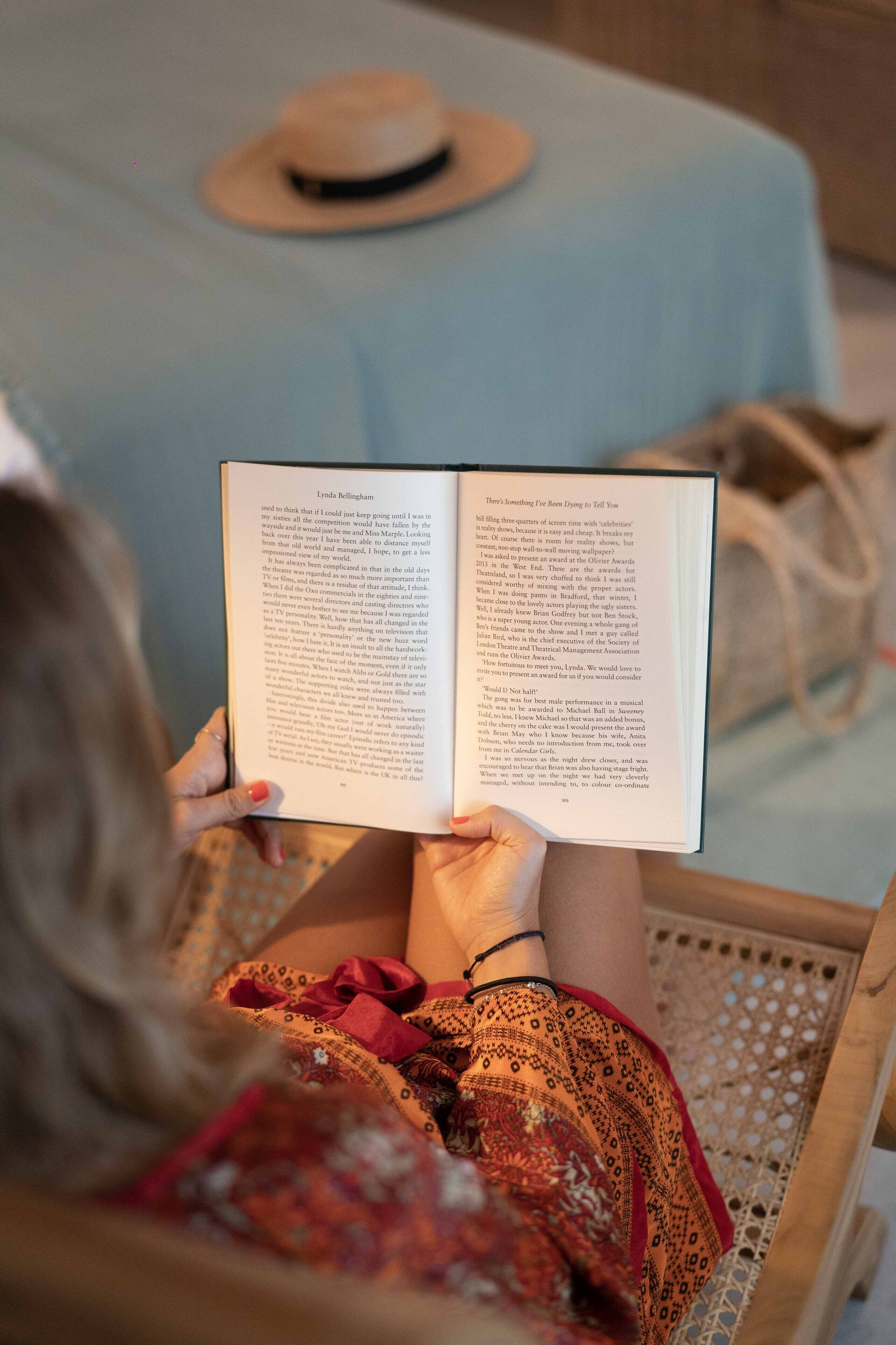 A person reading a book in a wicker chair with a light blue bedspread and a hat on the bed.