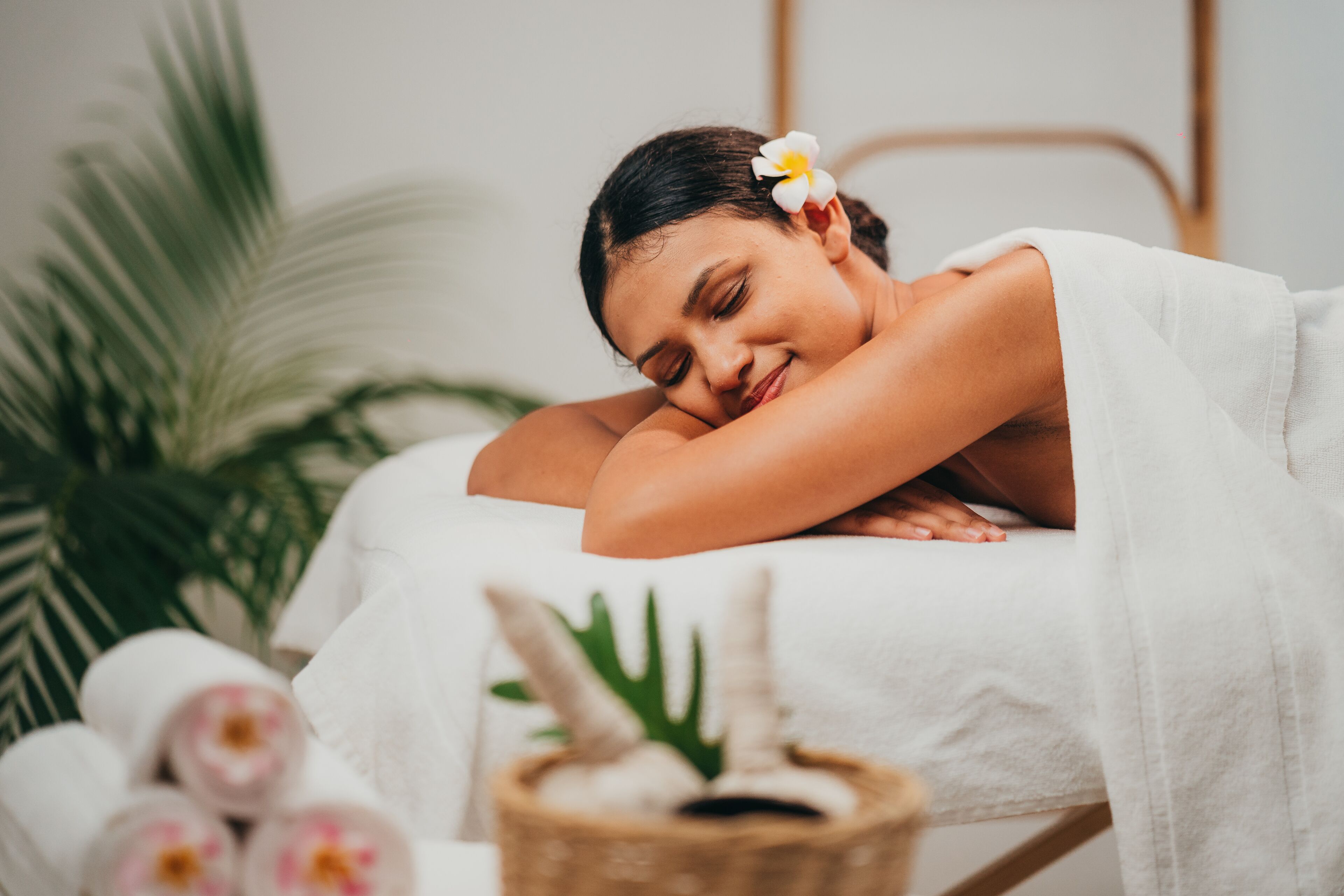 Una persona acostada en una cama con una toalla blanca, una flor en el cabello y una planta al fondo.