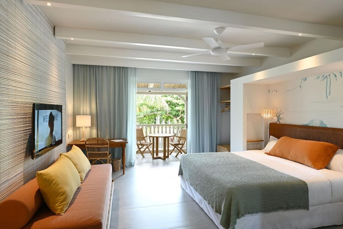 Una habitación de hotel con cama, sofá, televisor, balcón con mesa y sillas, y ventilador de techo.