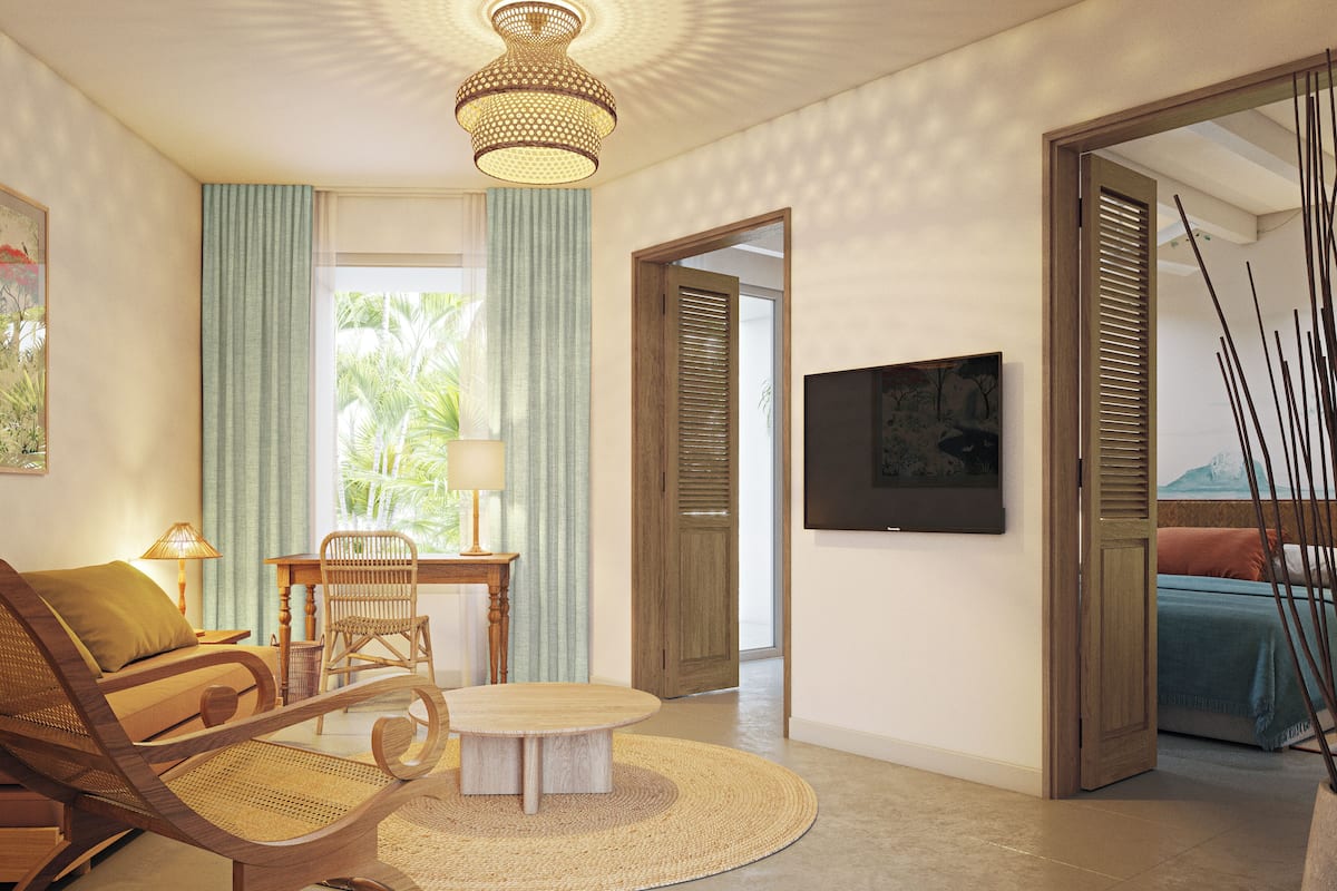 Una sala de estar bien iluminada con una mesa redonda de madera, una silla de mimbre y una mesa de comedor con sillas. Hay un televisor montado en la pared y una puerta que da a otra habitación.