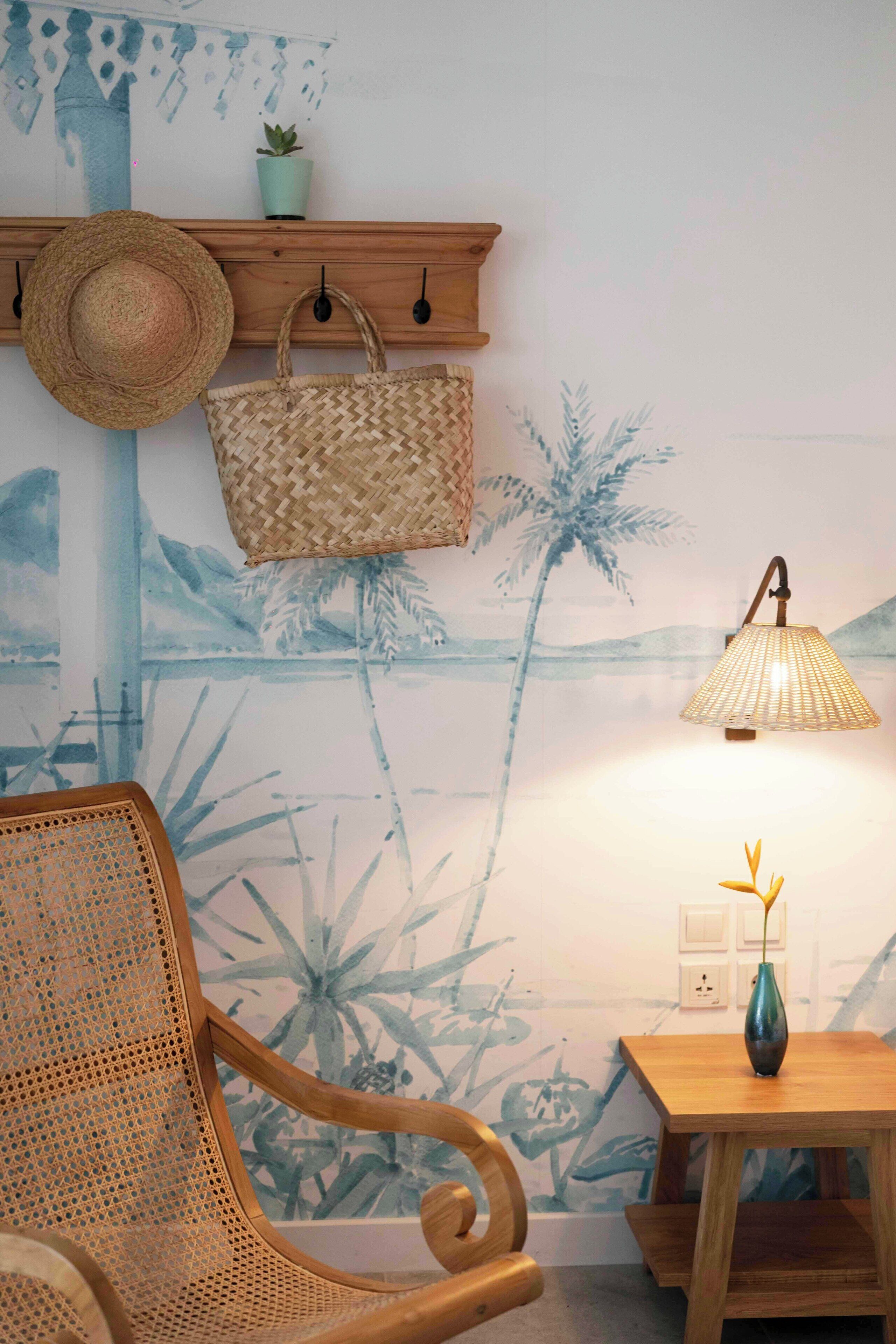 A room with a woven chair, a hanging hat, a woven bag, a small plant, and a lamp on a wooden table.
