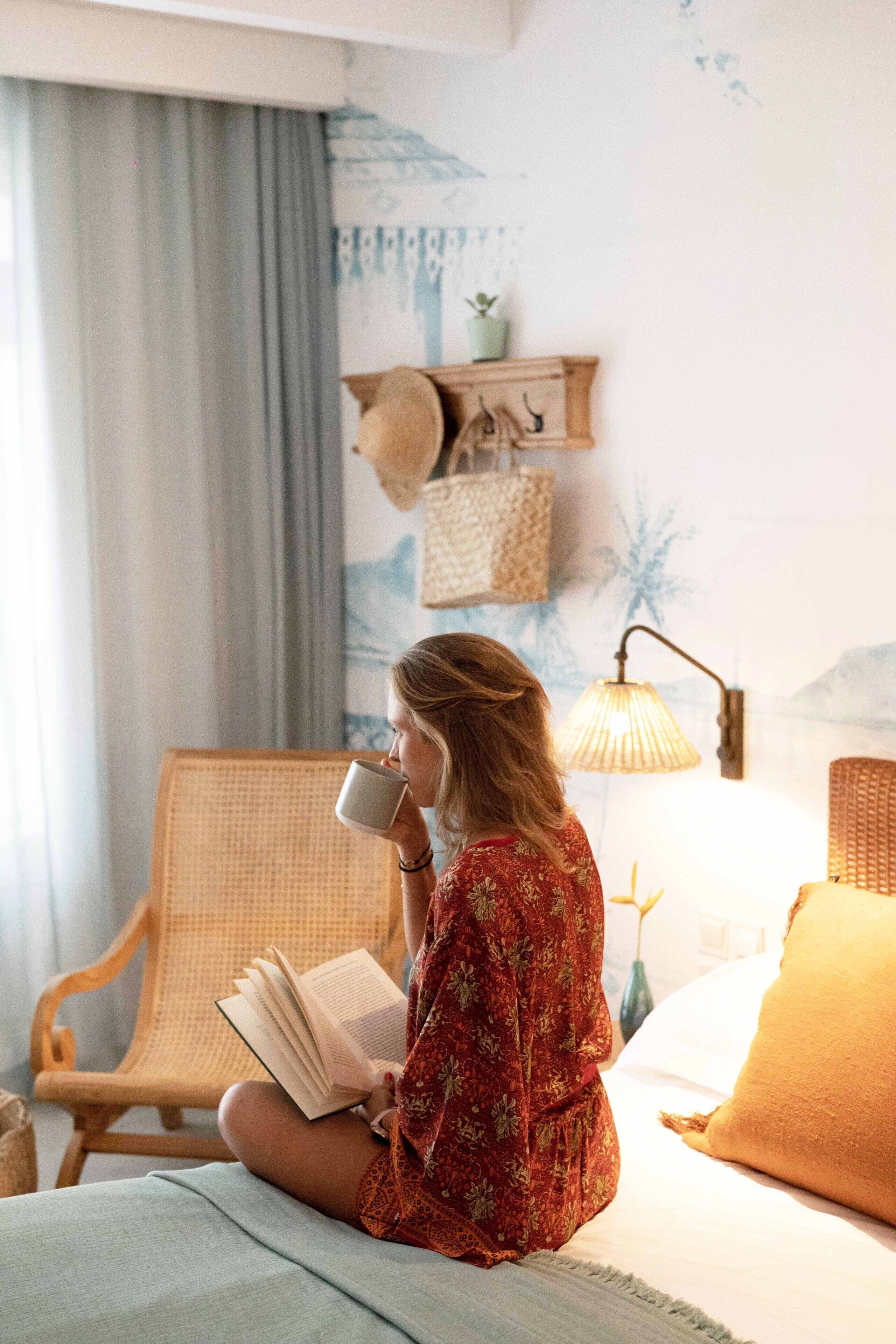 Une personne assise sur un lit, en train de lire un livre, avec une lampe et une étagère remplie de chapeaux en arrière-plan.
