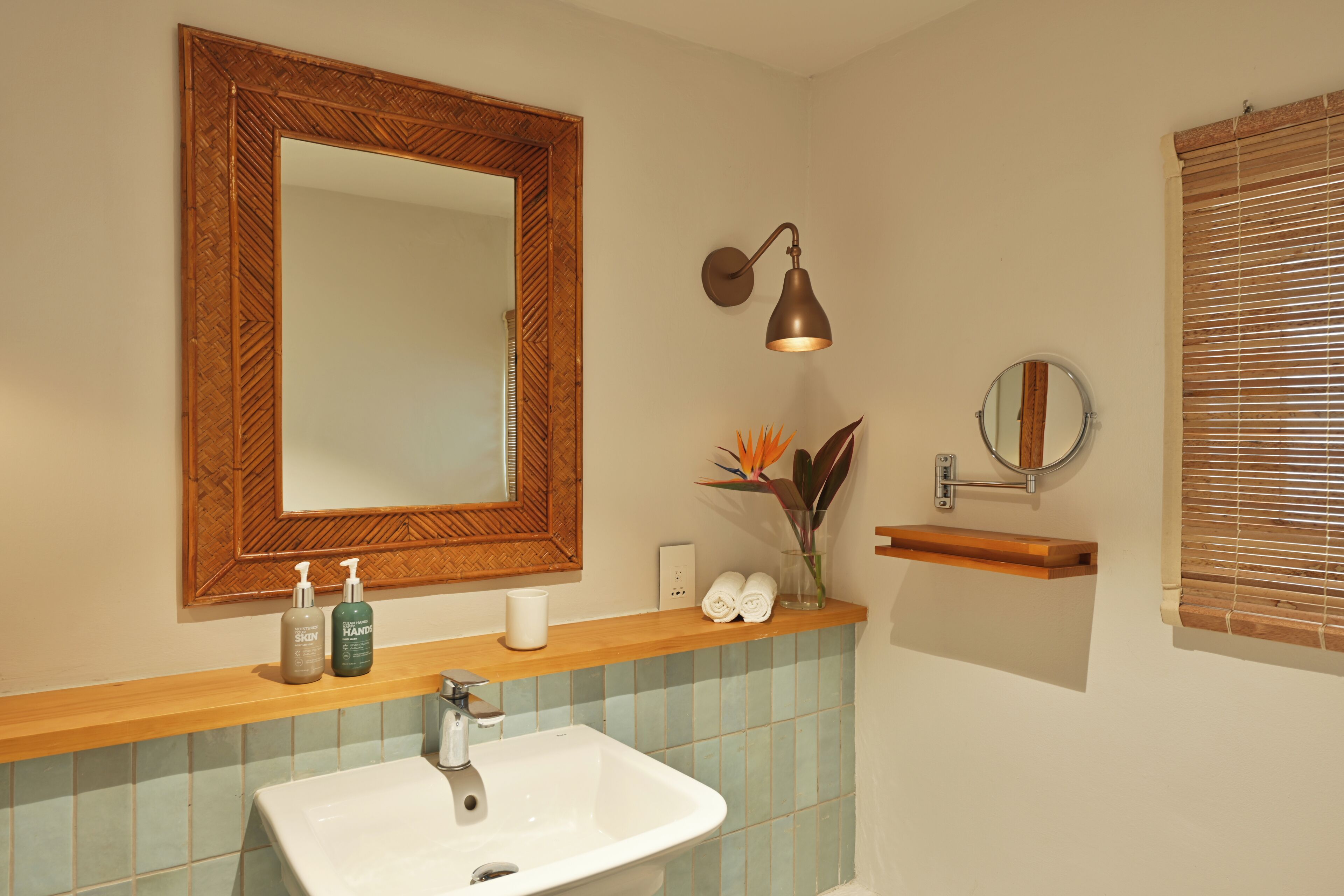 A bathroom with a wooden vanity, a white sink, a mirror, a wall-mounted lamp, and a window with blinds.