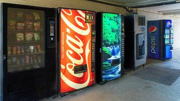 Una hilera de máquinas expendedoras con diversas marcas, incluyendo Coca-Cola, Pepsi, entre otras, con un muro de ladrillos al fondo.