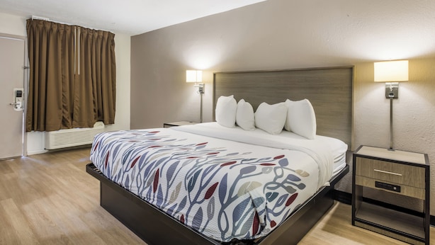Une chambre d’hôtel avec un grand lit, deux tables de chevet, une petite table, une lampe fixée au mur et une fenêtre avec des rideaux.