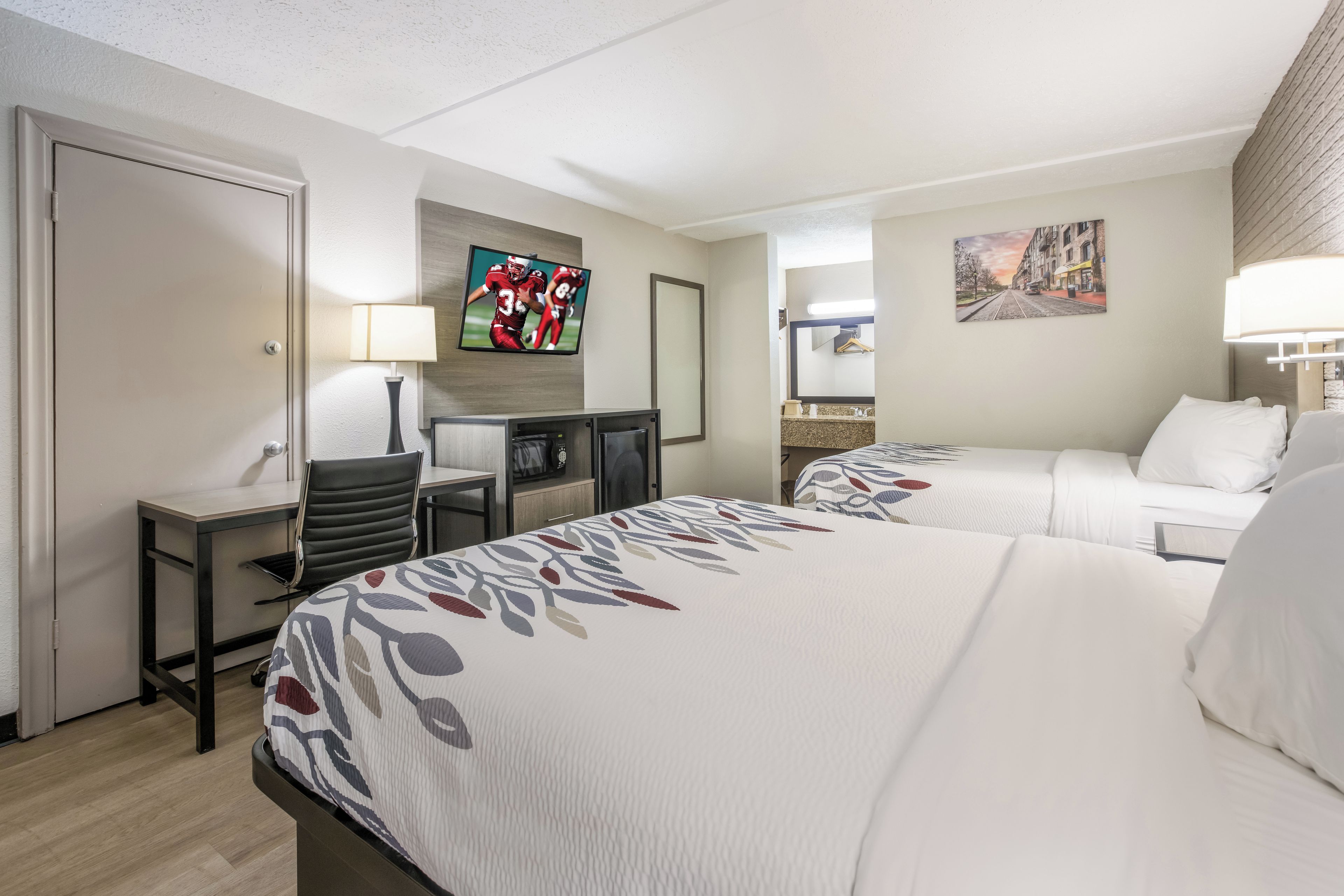 A hotel room with a bed, a desk, a chair, a television, and a painting on the wall.