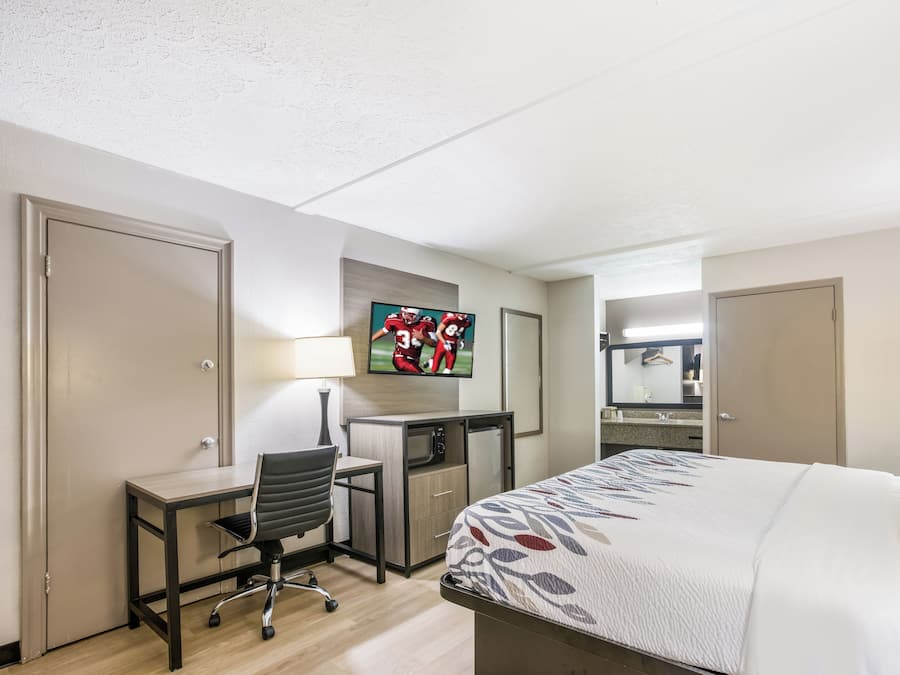 Habitación de hotel con cama, escritorio, silla, televisor y baño visible a través de una puerta abierta.