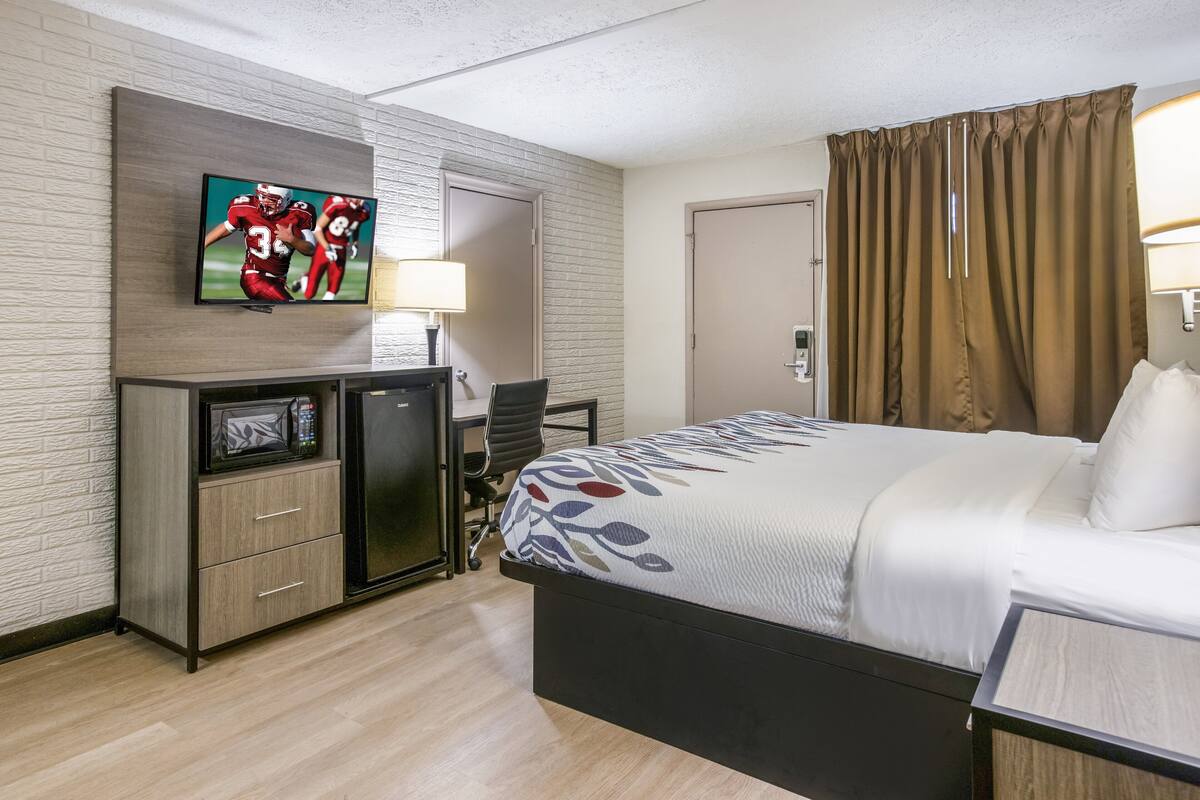 A hotel room with a bed, a desk with a computer, a television mounted on the wall, and a door with curtains.