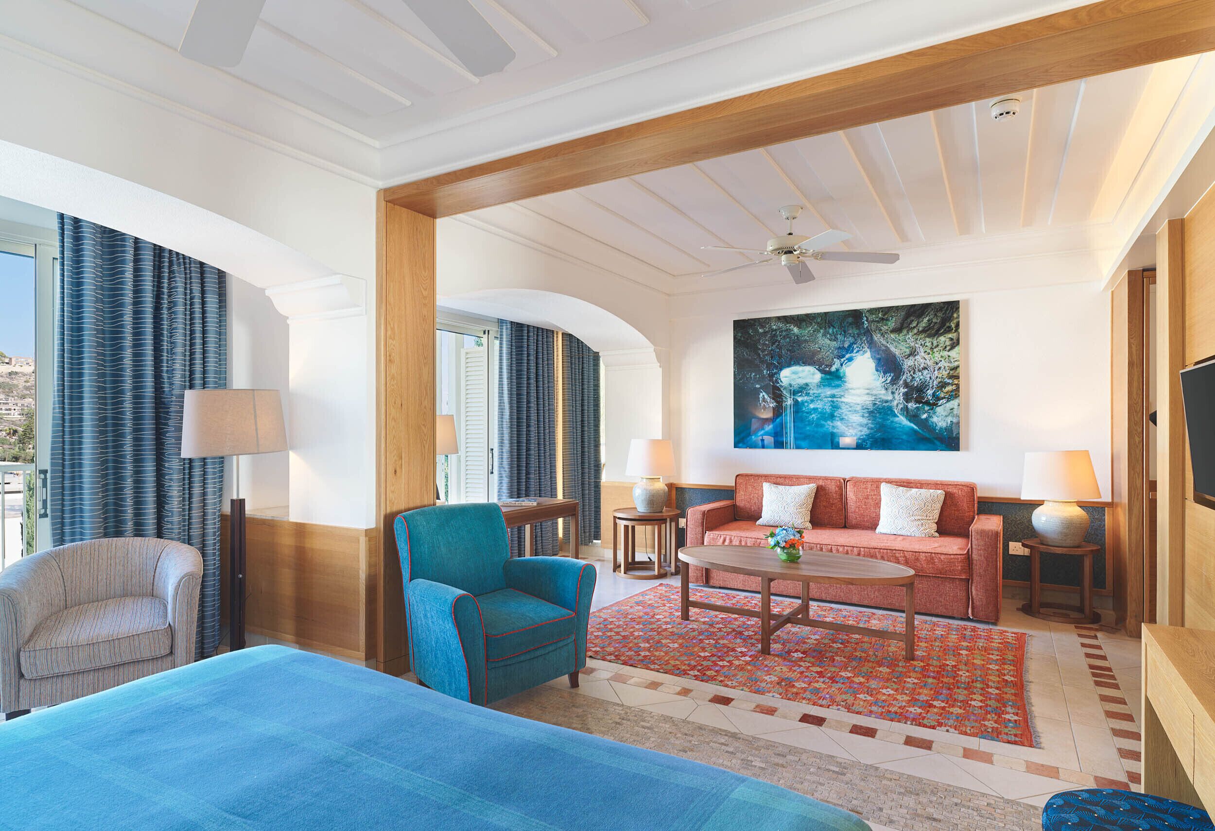 Una habitación de hotel con una cama, un sillón azul, un sofá rojo, una mesita de café y un cuadro en la pared.