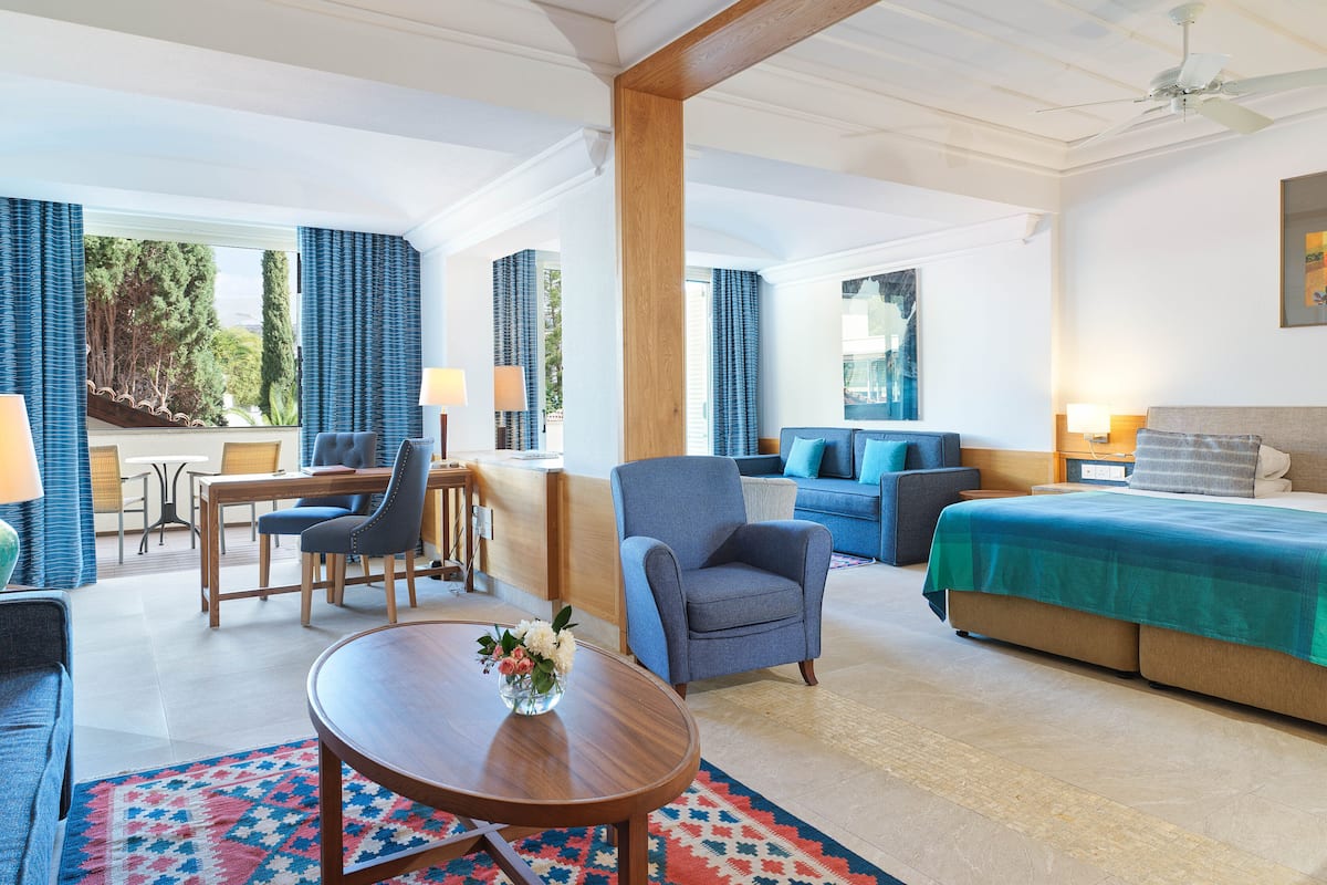 A hotel room with a bed, a sofa, a chair, a table, and a balcony with a view of trees.