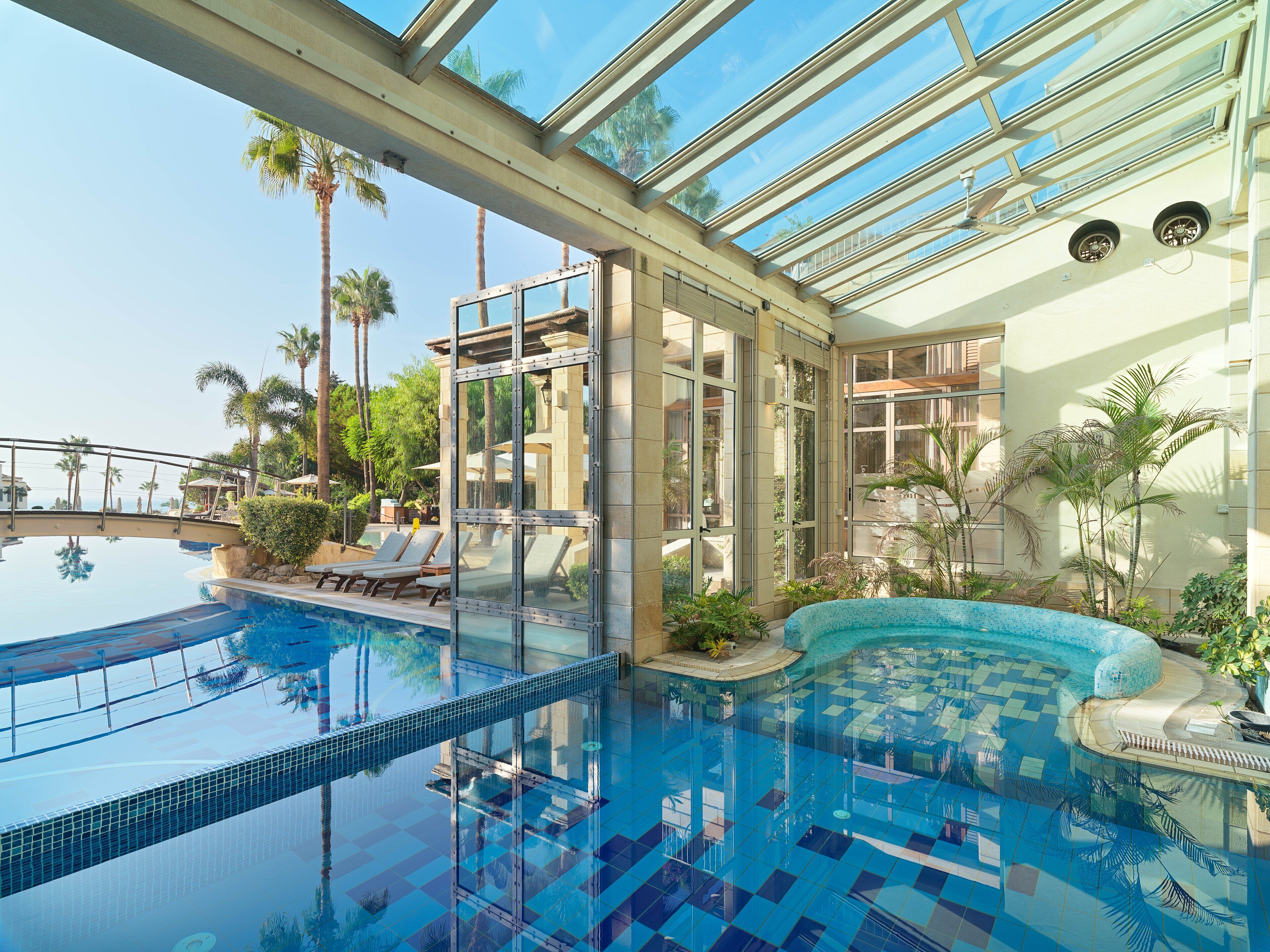 An indoor swimming pool with clear glass ceilings, lounge chairs, and palm trees.