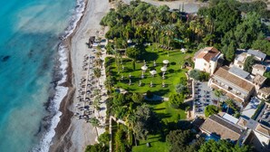 An aerial view of a coastal resort with a beach, palm trees, and a well-maintained lawn with statues. - Columbia Beach Resort (Pissouri)